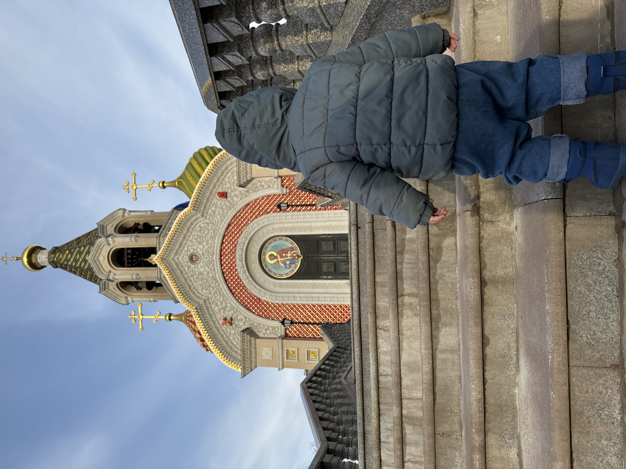 A child in a gray puffy jacket and blue pants is walking up stone steps. In the background is the Church of St. Igor Chernigovsky, located in Peredelkino, Moscow, Russia. The church is colorful and ornate, with multiple golden crosses on top. The sky is light blue with some clouds.