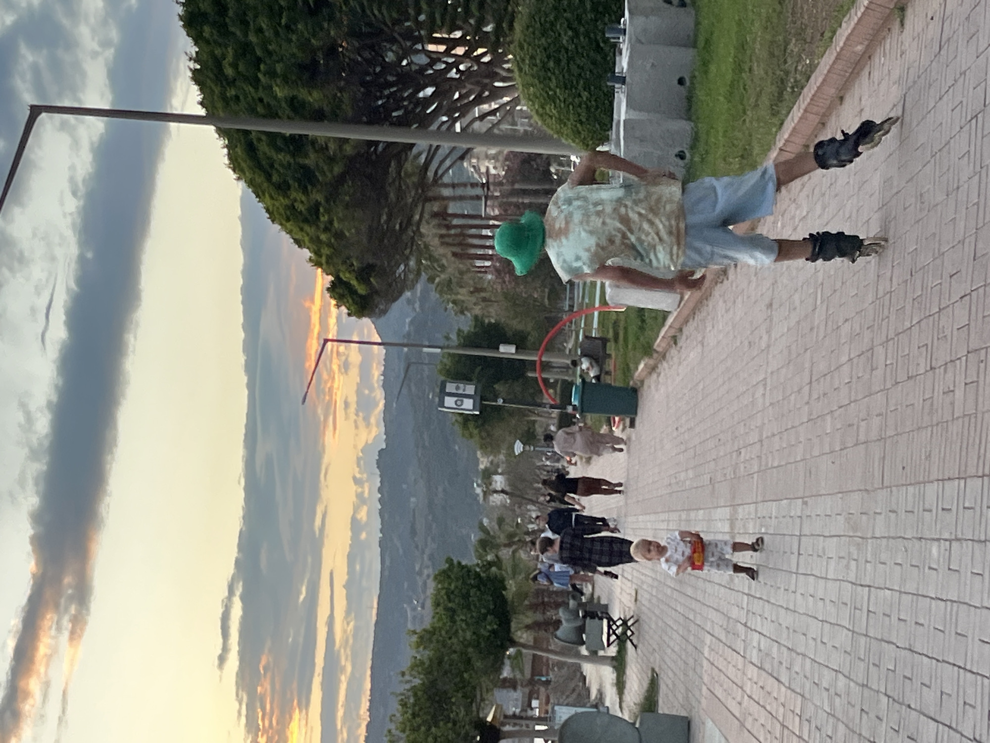 The image captures a scene along a pedestrian walkway in Alanya, Turkey. The sky shows a sunset with various shades of yellow, orange, and gray clouds. In the background, there are mountains and the sea.
On the walkway, several people are present, including a person roller-skating and another using a hula hoop. A child is also visible on the walkway, as well as other individuals strolling. The walkway itself is paved with rectangular blocks.
There are trees and green patches along the walkway, and streetlights can be seen in the vicinity. Some benches are also placed along the walkway. A mountain is visible in the distance.