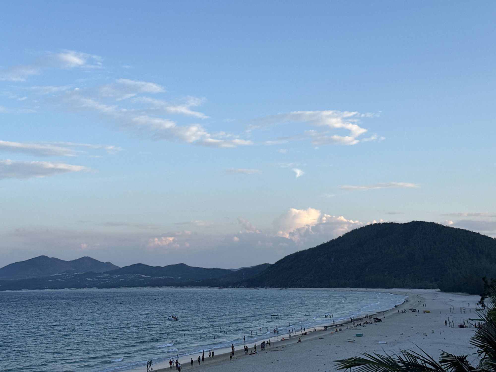 An aerial, eye-level, long shot captures a serene beach scene at dusk. The sky, a gradient of soft blues and pinks, is dotted with wispy white clouds. In the distance, a series of dark, undulating mountains hug the coastline. The ocean, a deep blue, meets the shore with gentle waves. A sandy beach stretches across the frame, populated by numerous beachgoers. People can be seen walking, swimming, and relaxing, some under colorful umbrellas. A small boat is visible on the water. Palm fronds frame the right side of the image, adding a tropical touch to the tranquil setting.