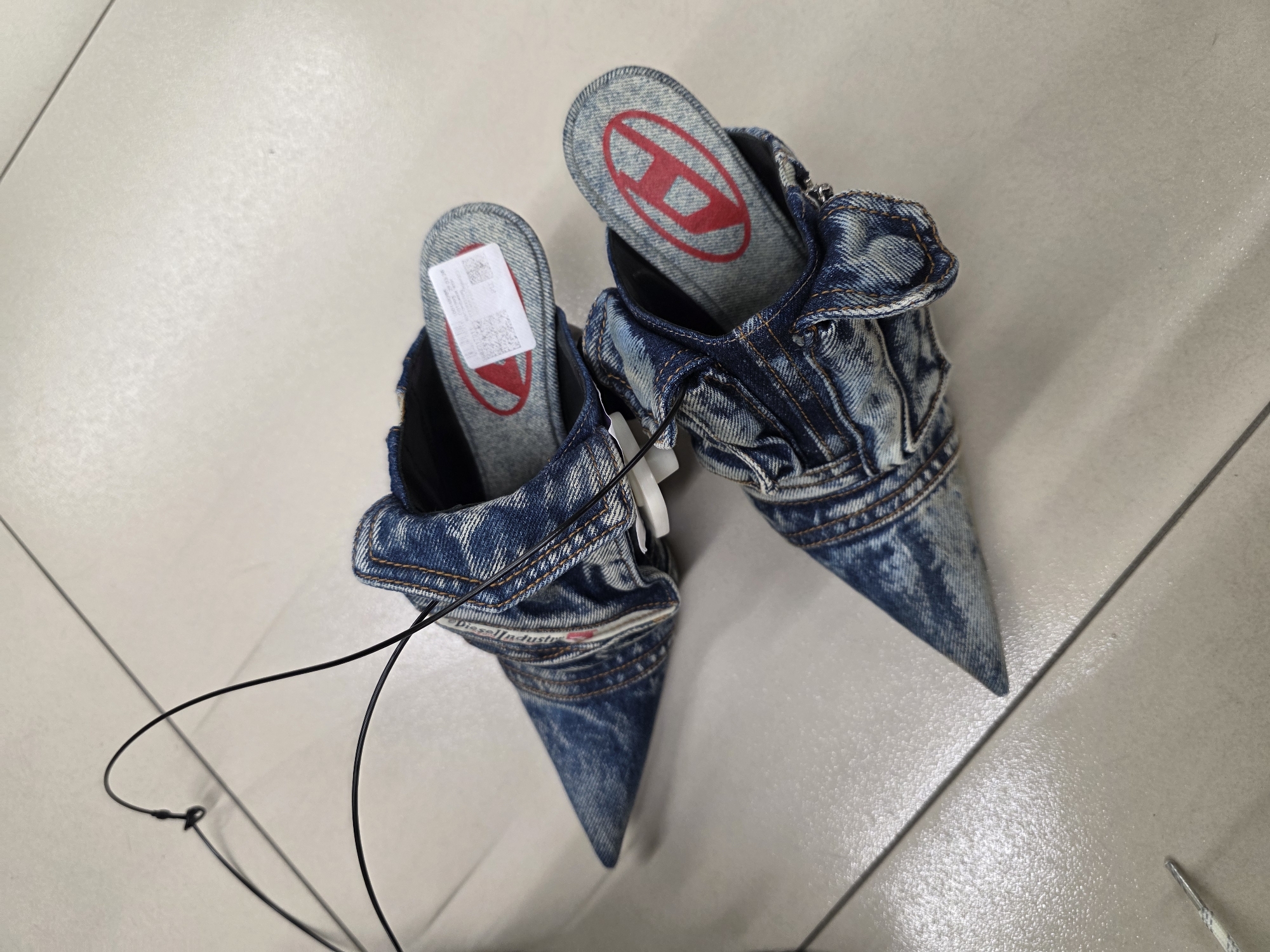 A pair of denim mules with pointed toes and red Diesel logos on the insoles are displayed on a light-colored tiled floor. The mules are made from distressed denim with visible stitching and texture. A black wire hanger is draped over the mules, with one end of the wire touching the floor near the toe of the mule in the foreground. The scene is photographed from a high-angle perspective. The floor tiles are arranged in a grid pattern with faint lines indicating grout. The lighting appears to be overhead and even. The overall environment suggests an indoor retail space. There are no people or animals visible in the image. The context of Saint Petersburg, Russia is not discernible from the visual elements of the image.
