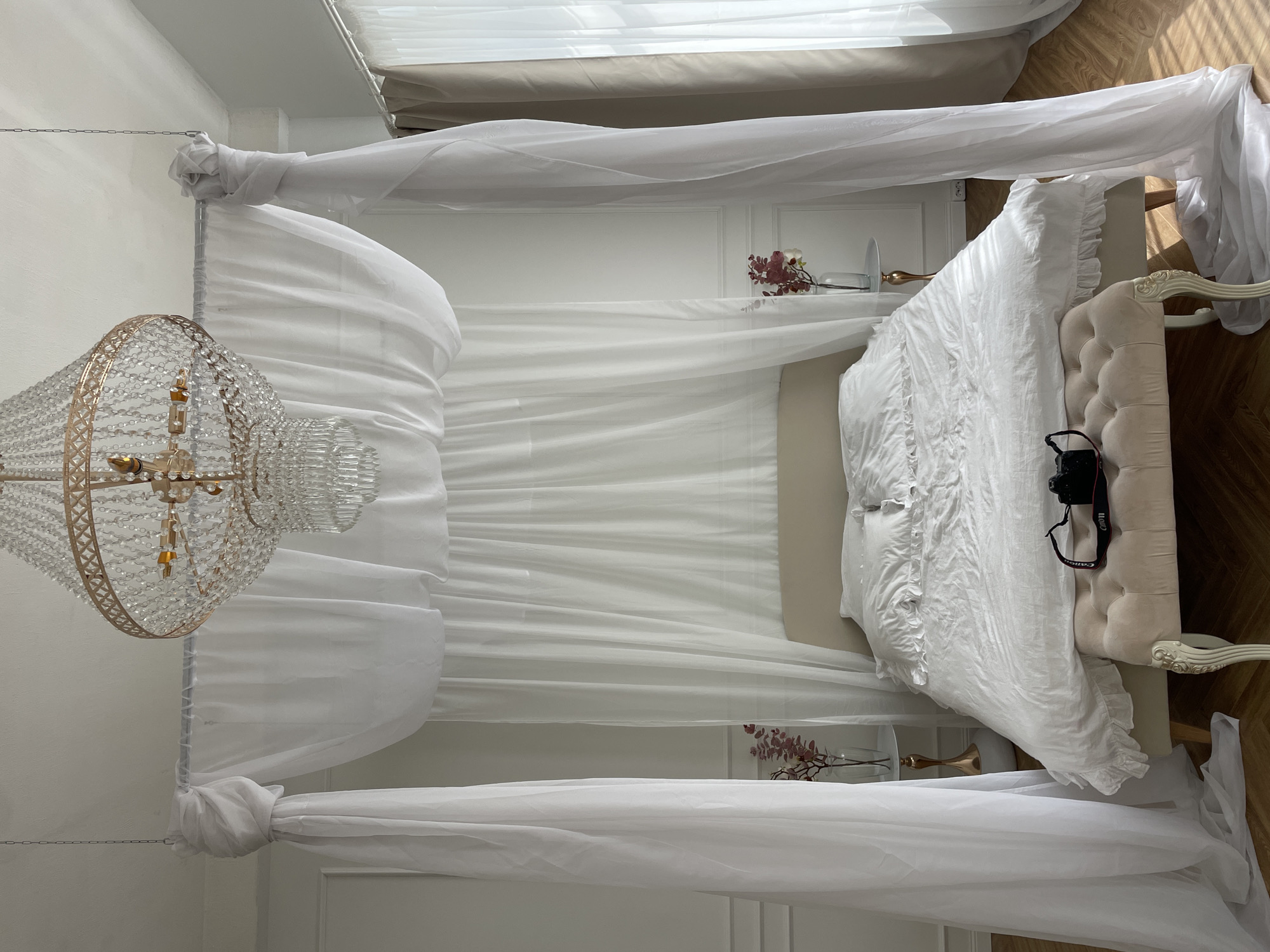 The image depicts a bedroom interior, likely staged for a photo shoot. The room features a four-poster bed draped in white sheer fabric, creating a canopy effect. A crystal chandelier hangs prominently to the left of the bed. The bed itself is made with white linens and a tufted headboard. A Canon camera is placed on the bed. The background includes white walls with decorative molding and a small vase of flowers on a bedside table. The floor appears to be hardwood. There are no people visible in the image.
The location is not discernible from the image itself, so stating it is in Kazan, Russia, would be speculative.