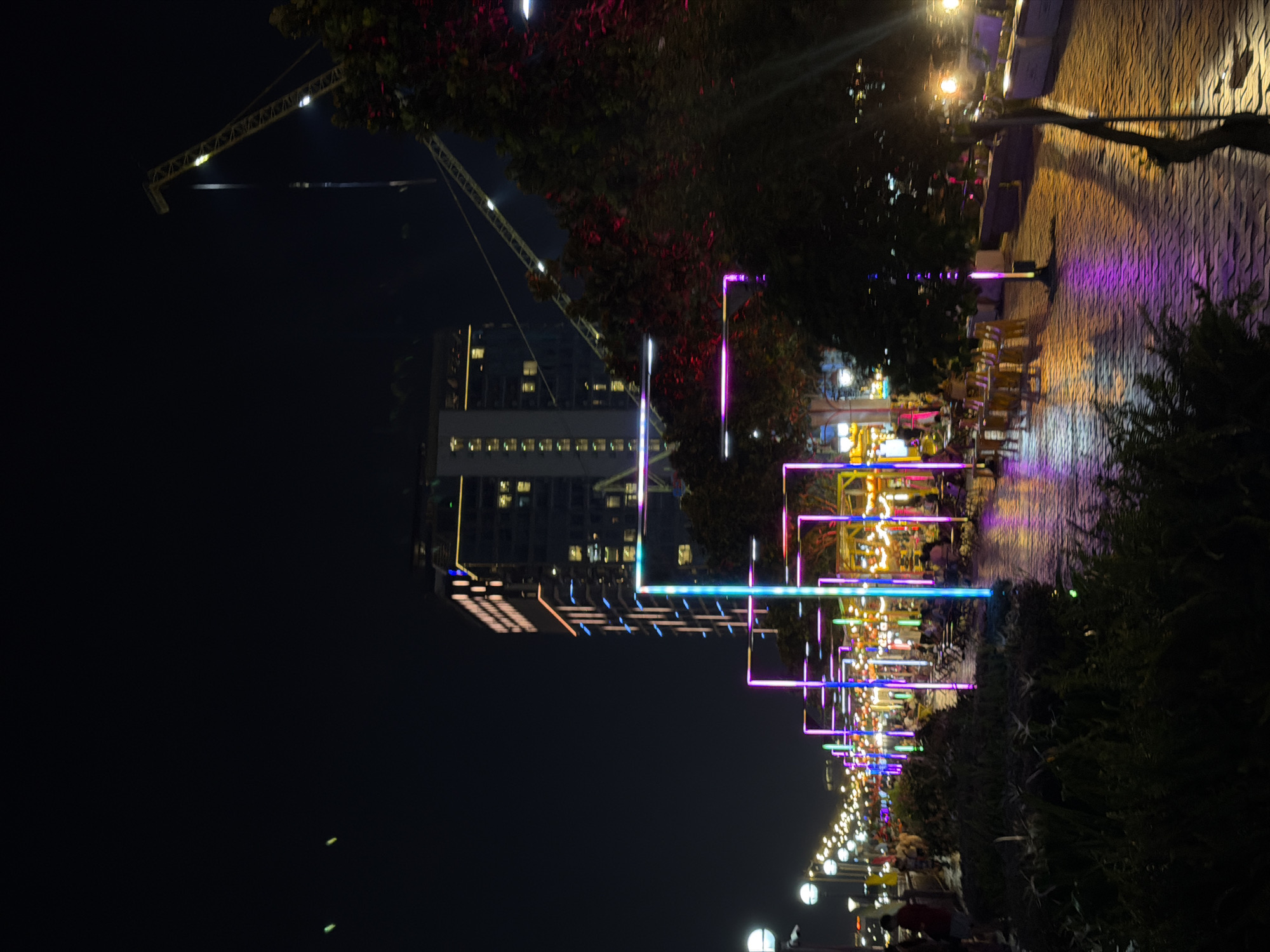 This nighttime photo captures a vibrant scene along a waterfront promenade in Đà Nẵng, Vietnam. The image is dominated by a building with lit windows in the background, suggesting an urban setting at night. A construction crane looms large in the upper left quadrant, hinting at ongoing development.
The foreground is illuminated by a striking display of colorful LED lights, forming abstract geometric patterns. These lights line what appears to be a pedestrian walkway, reflecting on a wet surface, possibly due to recent rain or proximity to water. The walkway is paved with textured cobblestones.
Scattered along the promenade are outdoor seating areas with tables and chairs, some of which are occupied by people. The overall atmosphere is festive and lively, with the bright lights creating a celebratory mood. The presence of people, outdoor dining setups, and the illuminated promenade suggests a popular gathering spot for locals and tourists alike.
The time of day is clearly night, and the reflections on the ground indicate a possibly damp or humid environment. The context of Đà Nẵng, Vietnam, combined with the modern architecture and the colorful light installations, points to a contemporary urban waterfront experience. No visible text is present in the image.