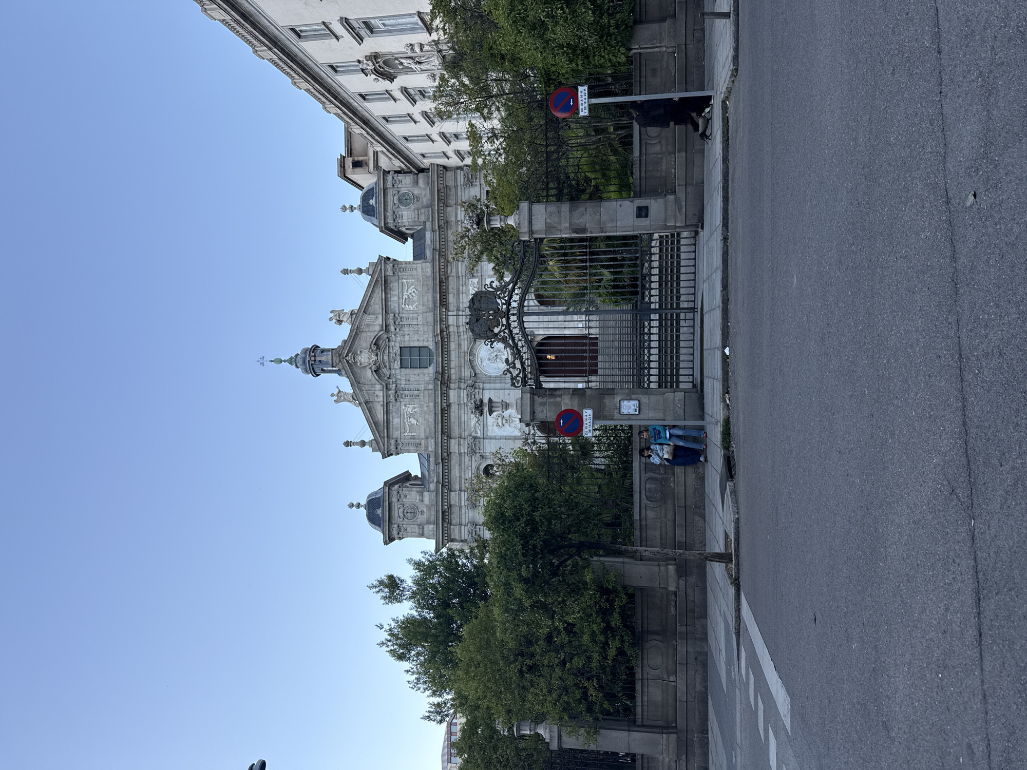 The image shows the Iglesia de San José in Madrid, Spain. The church is a baroque-style building with an ornate facade and a metal gate in front. A street runs along the side of the church, and a pedestrian crosswalk is visible. A parking restriction sign is displayed near the entrance. Two individuals stand on the sidewalk adjacent to the church. The sky is a clear, light blue. Another building of similar construction is to the left of the church.