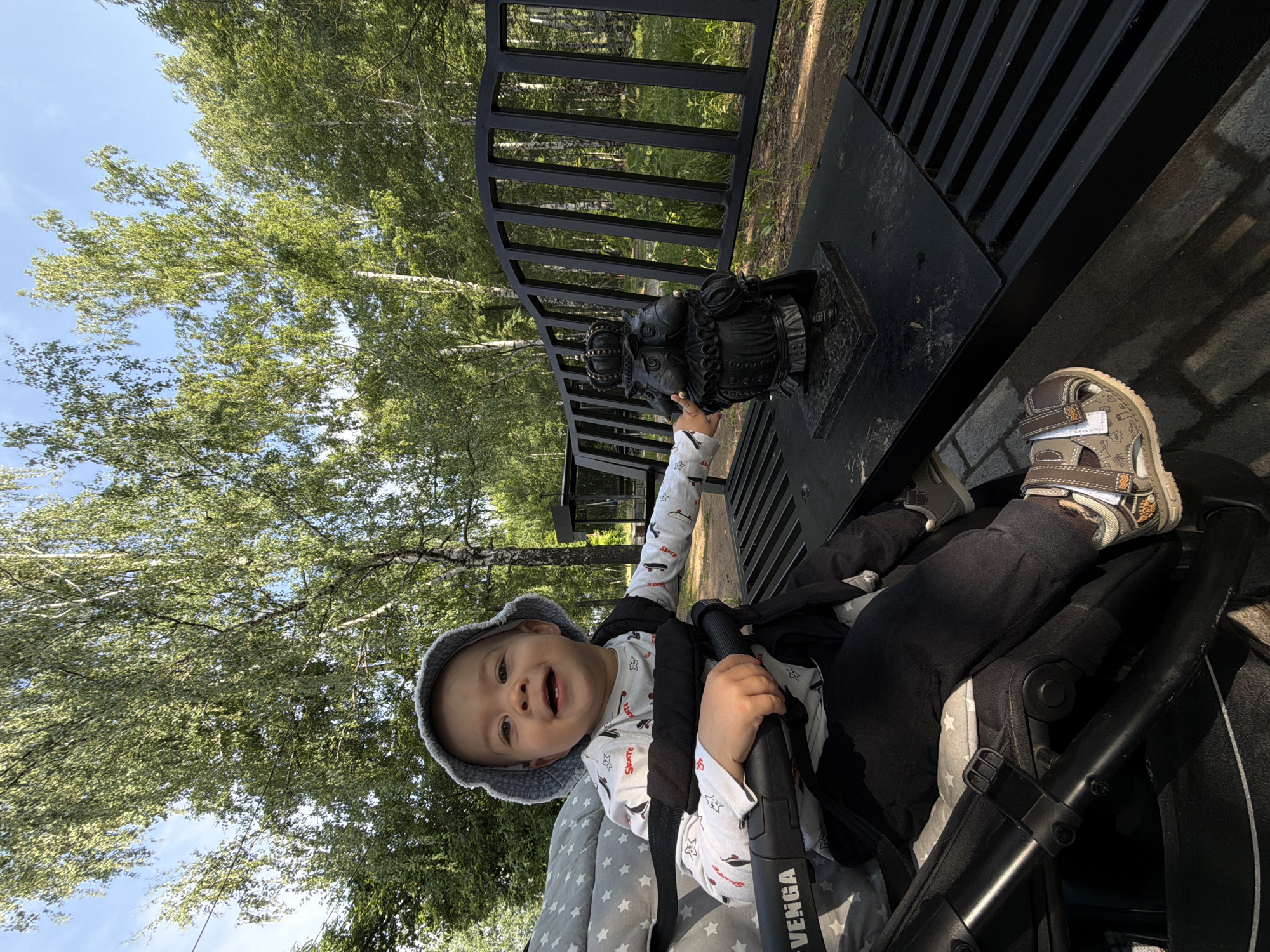 The image shows a baby sitting in a stroller in a park. The baby is wearing a light blue bucket hat and a white long-sleeved shirt with a skateboard print. The baby is smiling and looking at the camera. The stroller is black with white stars on the fabric. To the right of the stroller, there is a black bench with a small, dark statue of a figure wearing a crown. The background consists of trees with green leaves and a blue sky. The bench and statue are on a paved area, suggesting a park or public space. The overall scene suggests a pleasant day outdoors.