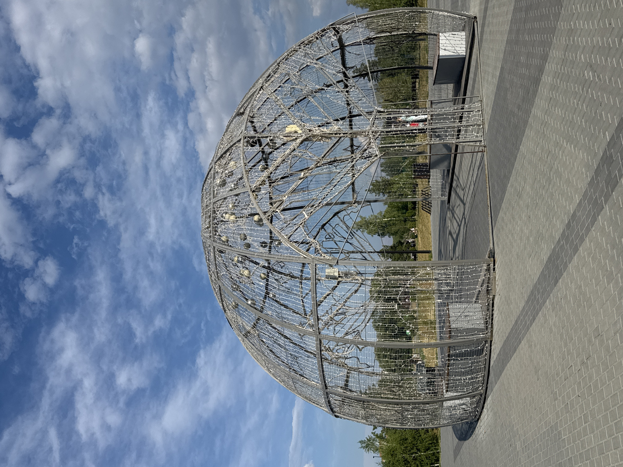 An exterior, full-shot photograph captures a large, geodesic dome-like structure adorned with numerous small lights, resembling a wireframe globe. The dome is constructed of metal framework and appears to be a decorative installation. Inside the dome, there are benches and what appears to be an internal structure with branches, also outlined with lights. The dome is situated on a paved area made of gray rectangular bricks. In the background, trees and a partly cloudy blue sky are visible. The scene suggests an outdoor public space, possibly a park or a recreational area. The structure is located in the city of Novosibirsk, Russia.