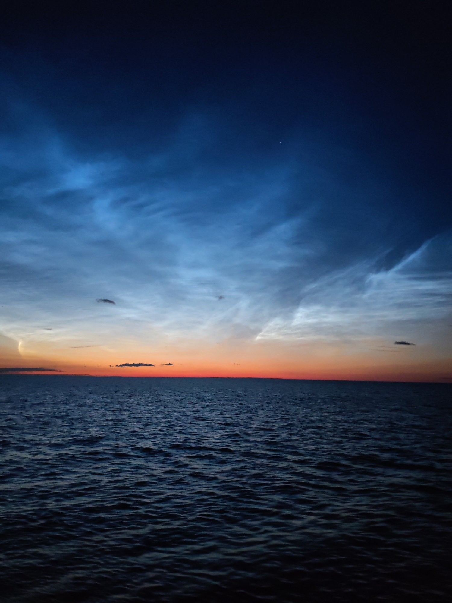 An image captured at twilight shows a serene body of water, likely an ocean or large lake, reflecting the sky above. The water's surface is textured with gentle waves, appearing dark blue and almost black in the low light. The sky is a gradient of colors, transitioning from a deep, dark blue at the top to lighter blues and oranges near the horizon. Wispy, luminous clouds are scattered across the sky, catching the last rays of sunlight and appearing almost ethereal. A thin sliver of orange or yellow is visible at the horizon, marking the sunset. The scene is devoid of any human presence or man-made structures. The location is stated as Unknown, Russia.
