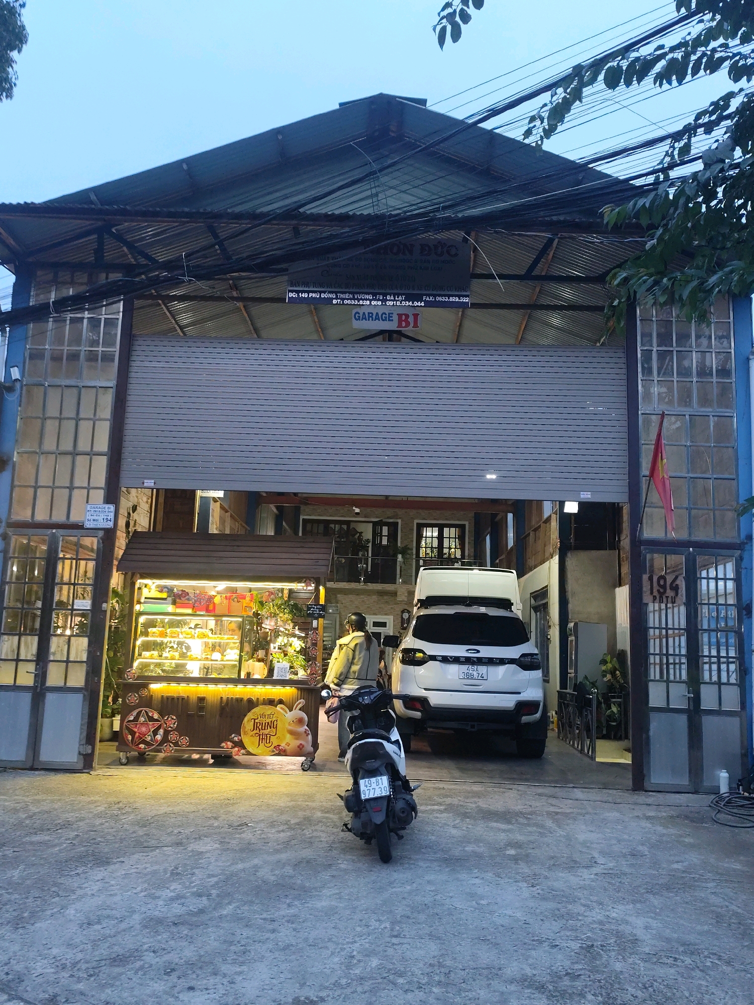 In the city of Lâm Viên - Đà Lạt, Vietnam, this image captures a street-level view of a garage and a food stall. The building has a large, corrugated metal roll-up door, likely for the garage. To the left of the garage door, there are large windows with many panes, some of which appear to be industrial or salvaged. A smaller, more ornate window structure is visible below these. A food stall with illuminated display cases is set up to the left of the garage entrance, featuring pastries and decorative items, including a cartoon rabbit holding a sign that reads "Vui Tết TRUNG THU." A dark-colored motorcycle with a license plate (49-B1 977.39) is parked in the foreground, facing away from the camera. A white SUV with a license plate (49A 366.74) is parked inside the garage entrance. A person wearing a light-colored jacket and helmet stands near the food stall, facing away from the camera. A red Vietnamese flag is mounted on a pole to the right of the garage door. The overall scene suggests a commercial area, possibly with residential units above. The sky is overcast and the lighting indicates either early morning or late evening.