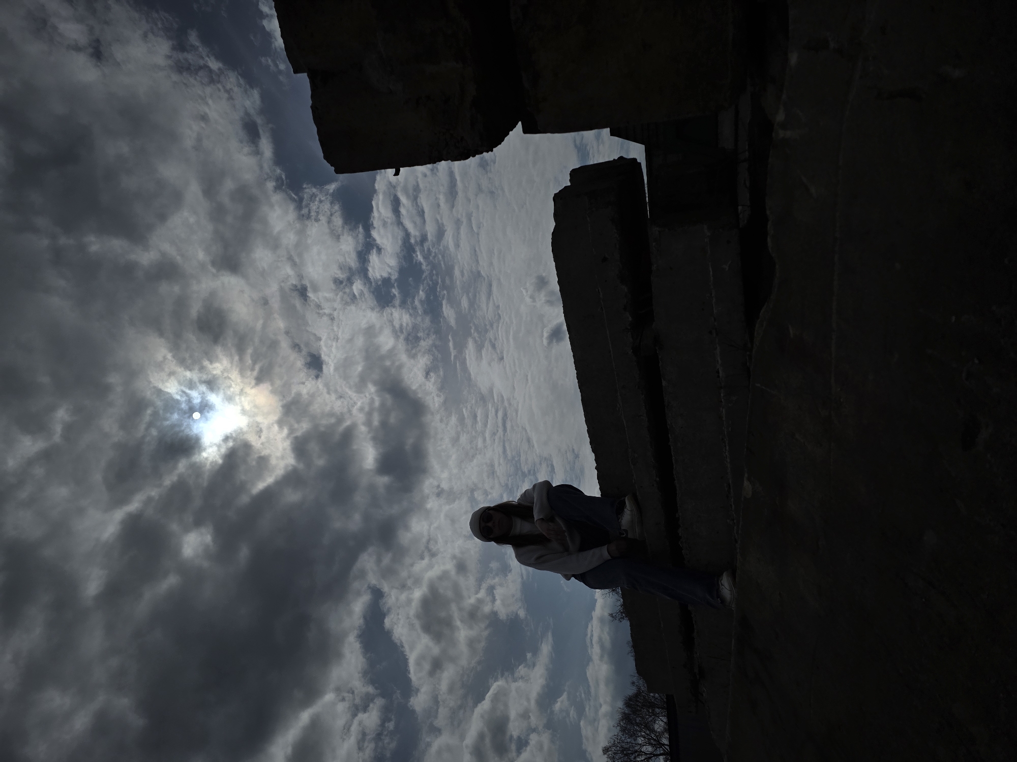A full shot, rotated 90 degrees to the left, presents a woman in jeans, white shoes, and a light-colored top, perched on a dark, weathered structure, possibly concrete or stone. The structure appears to be a fragment of a building or wall. The sky behind the woman features a mix of light and dark clouds, with the sun shining through. Some barren tree branches are seen at the bottom edge of the frame. The location appears to be in a ruined or abandoned area of a city. The woman is looking directly at the camera, but with the image rotation, she appears to be seated upright on a vertical surface.