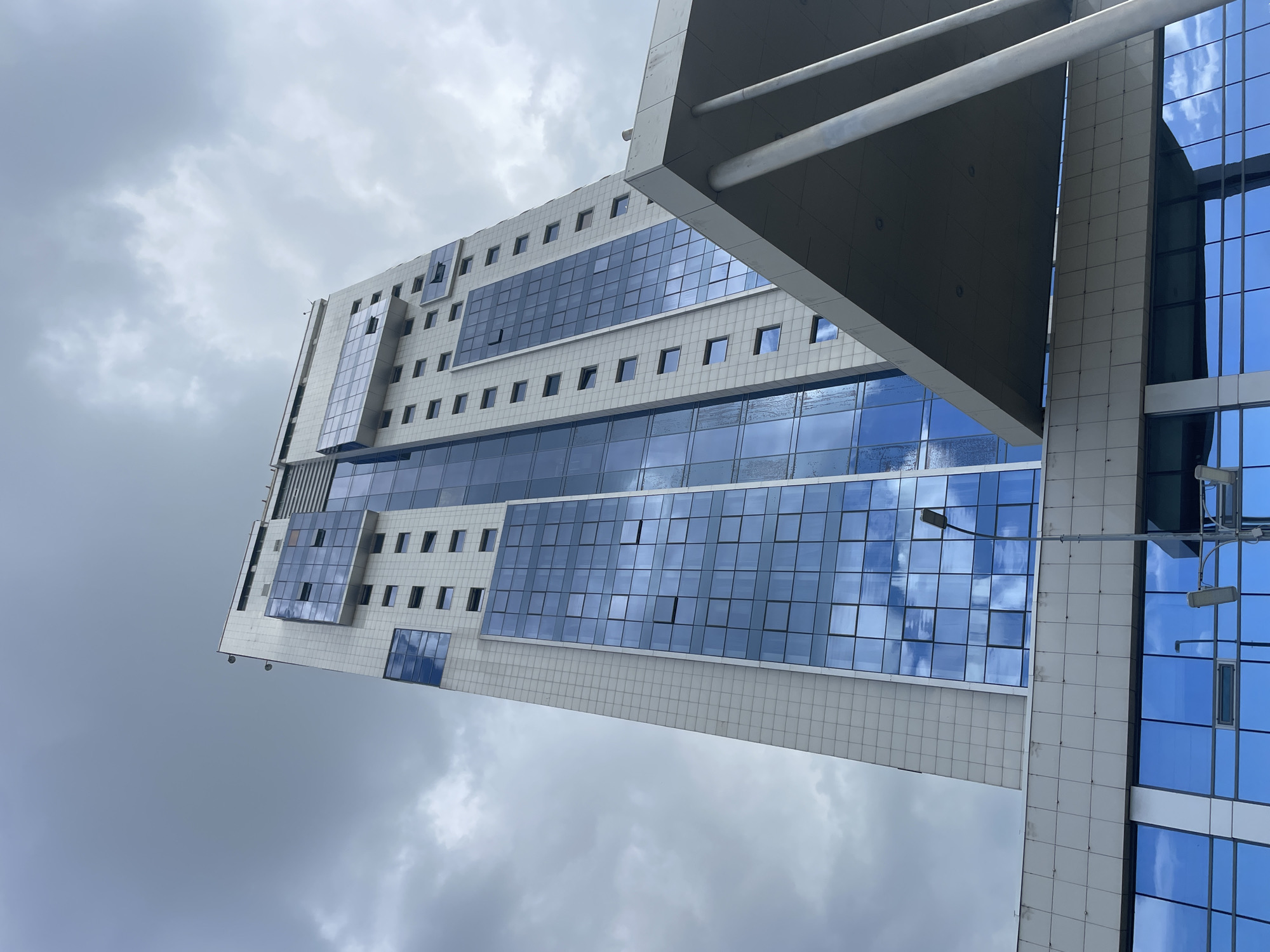 The photograph captures a low-angle view of a modern skyscraper in Vysokaya Gora, Russia. The building features a white facade interspersed with large, blue-tinted glass windows that reflect the overcast sky. The architecture is characterized by sharp, geometric lines and protruding sections with glass panels. The lower part of the building exhibits a more horizontal design with a darker band of blue-tinted windows. A cantilevered section with thick support columns extends from the building in the foreground. A street lamp is visible to the left of the skyscraper, and traffic lights are also present in the lower portion of the image. The sky is filled with heavy, gray clouds, suggesting a cloudy or pre-stormy day. No people are visible in the frame.