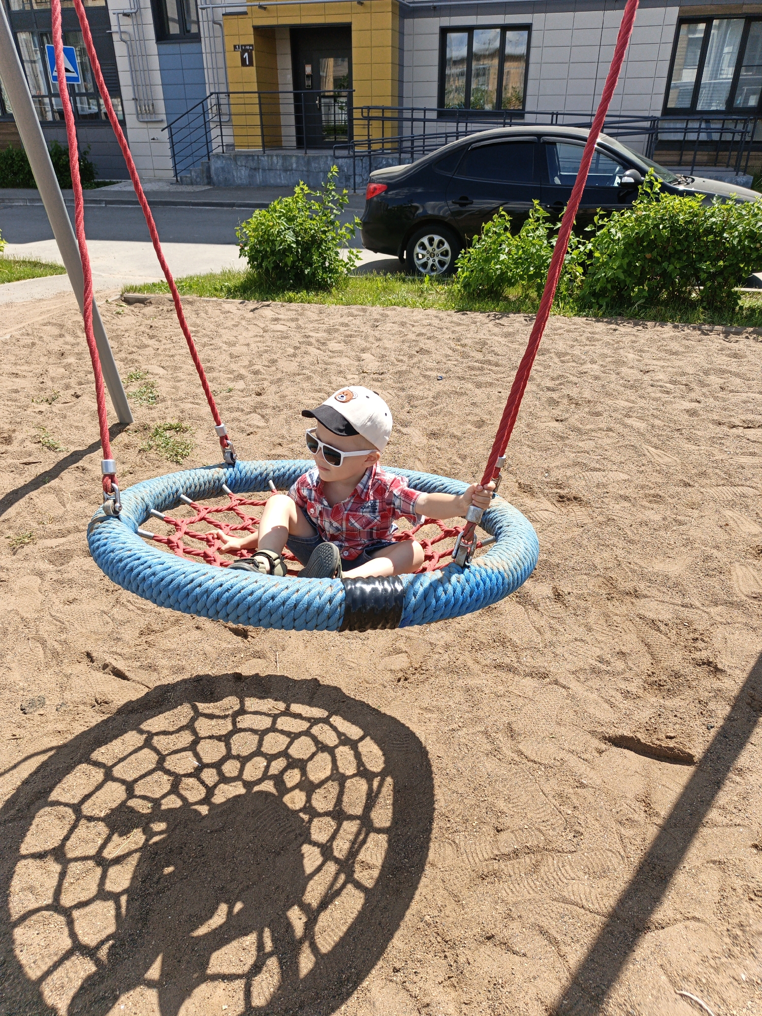 A young boy sits on a round, blue rope swing in a sandbox. He is wearing a red and white plaid shirt, blue shorts, a baseball cap, and white sunglasses. The swing is suspended by red ropes from a metal frame. In the background, there is a modern building with yellow and grey facades, and a black car parked on the street. The scene appears to be a residential area in Kuyuki, Russia.