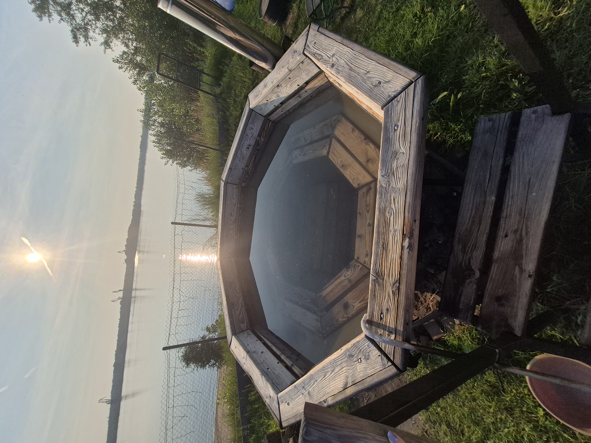 The image shows an octagonal wooden hot tub filled with dark water, situated outdoors on a grassy area near a body of water. A metal chimney pipe extends upwards from the hot tub, suggesting it is heated. Wooden planks form a small bench or seating area to the right of the hot tub. In the background, a calm lake or river stretches out, with a distant shoreline visible. The sun is setting or rising, casting a warm glow and creating reflections on the water. The overall scene evokes a sense of relaxation and tranquility. There is no visible text in the image.