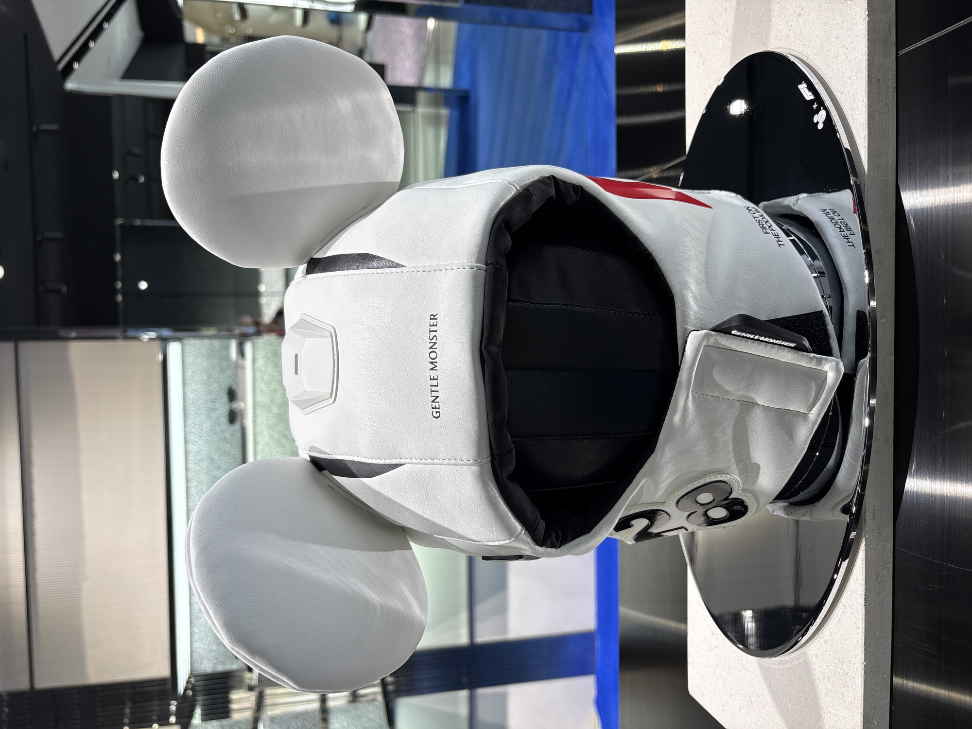 A white helmet resembling Mickey Mouse ears sits on a reflective black stand. The helmet has the words "GENTLE MONSTER" printed vertically on its face. The number "28" is visible on the right side of the helmet, near the stand. The location appears to be a store or display area in Bangkok, Thailand, as the image was generated using that information. The helmet is the central object.
