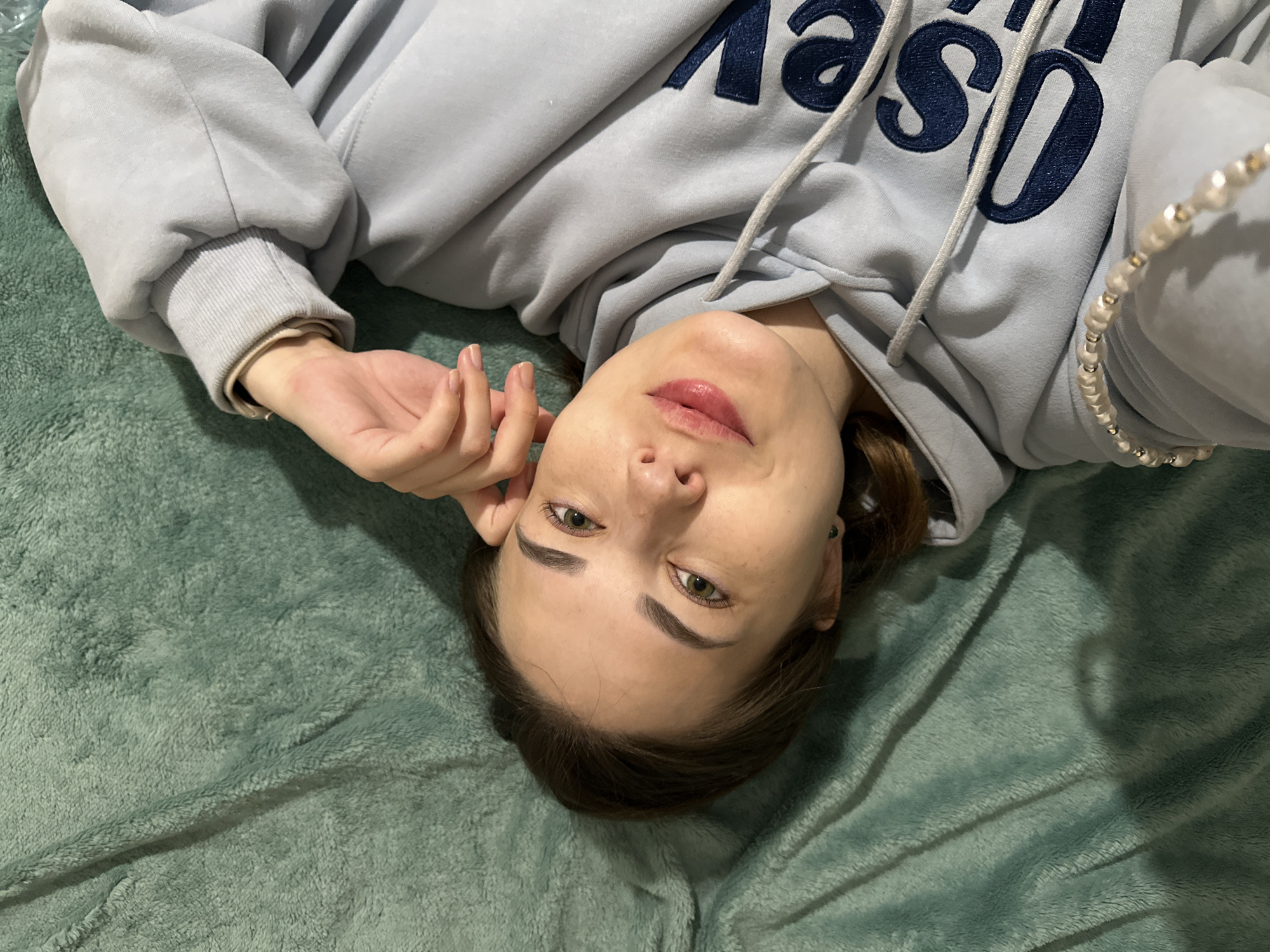 A young woman is depicted lying on her back, looking directly at the camera with her head positioned towards the bottom of the frame, creating an inverted perspective. She has green eyes, dark eyebrows, and dark brown hair. Her lips are adorned with pink lipstick, and her expression is calm and relaxed.
She is wearing a light blue or pale grey hooded sweatshirt (hoodie) that features dark blue lettering across the chest. The letters "OS" are clearly visible, suggesting part of a word like "ODYSSEY," along with white drawstrings around the hood. A beaded bracelet, appearing to be made of pearl-like beads interspersed with small silver accents, is worn on her right wrist.
The woman is resting on a plush, textured teal or dark green blanket or bedspread, which forms the background of the image. The setting is indoors, with even, artificial lighting. The scene captures a casual and personal moment. The location of the photo is Perm, Russia.