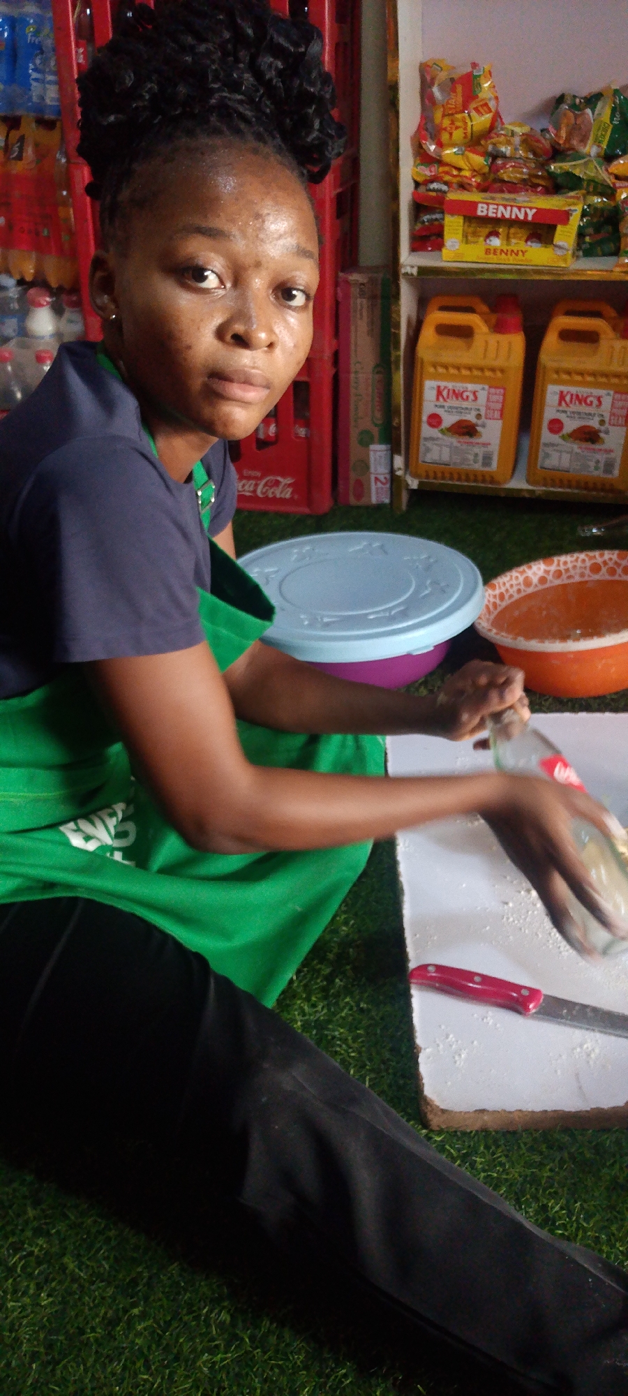 A woman is kneeling on a patch of artificial green grass in what appears to be a small shop in Adankolo, Nigeria. She is wearing a dark blue shirt, a green apron, and black pants. Her hair is styled in a high bun. She is rolling a Coca-Cola bottle on a white surface that is covered with a white powdery substance; a knife with a red handle is beside the board. To the right of the board is an orange bowl. Behind her, there's a shelf containing various products, including cases of Coca-Cola, jars of vegetable oil, and other food items.