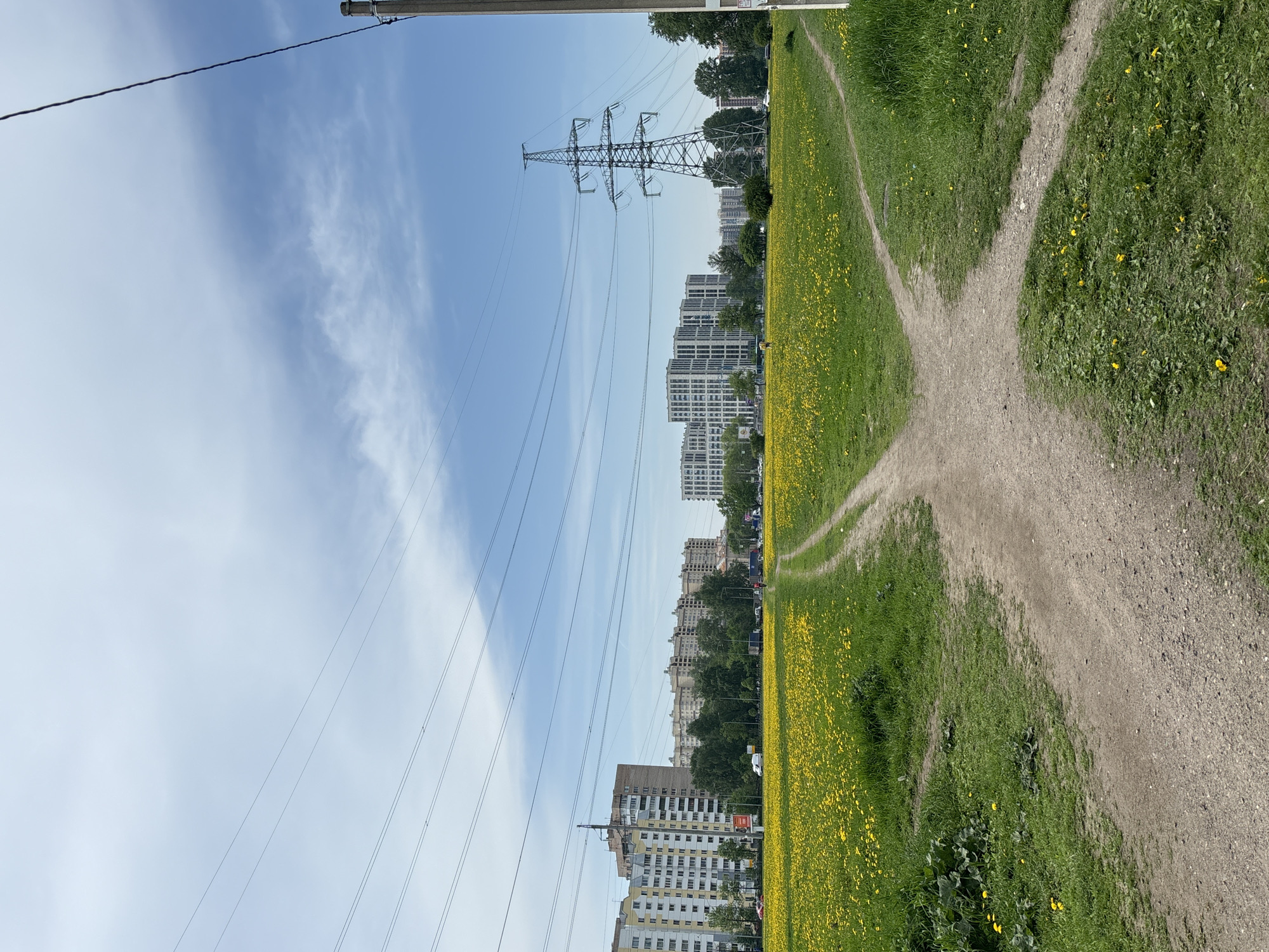The image depicts an outdoor scene in an urban area. In the foreground, a dirt path intersects with two other paths, creating a junction. The paths are surrounded by green grass with numerous yellow flowers scattered throughout. In the background, several high-rise buildings are visible, with an electrical transmission tower positioned near them. Power lines stretch across the sky from the tower towards the lower part of the frame. The sky is blue with some white clouds. There are no people visible in the image. The location appears to be a park or green space within a city.