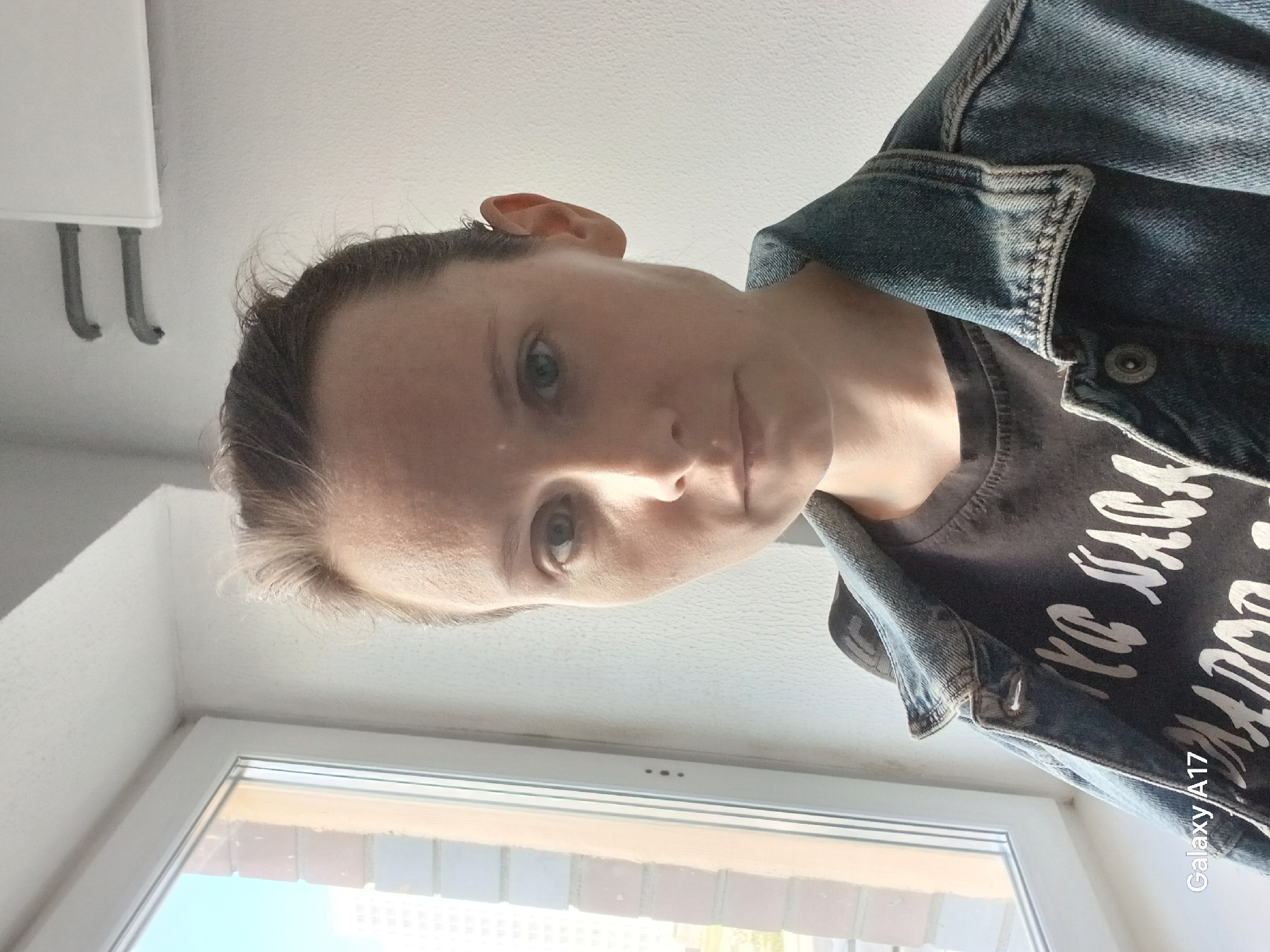 A Caucasian woman is depicted in an indoor setting, possibly a residential space. She is wearing a denim jacket over a printed gray t-shirt. Her hair is pulled back, revealing her face. The background includes a window showing a view of what appears to be the city of Yekaterinburg, Russia. The window frame and the adjacent wall are white. To the top left, there are some plumbing fixtures visible. The image is captured at an upward angle. A "Galaxy A17" watermark is visible on the lower right.