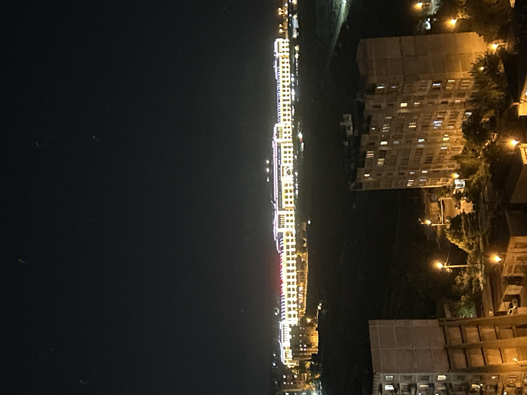 An aerial, nighttime photograph captures a wide, long, horizontally oriented building illuminated with bright white and red lights, stretching across the middle ground. The building appears to be a commercial or public structure, possibly a shopping mall or a large administrative complex, with numerous windows visible along its facade. In the foreground, two residential buildings are visible; the one on the left is a tall, concrete structure with visible windows lit from within, while the building on the right is also a multi-story residential building, appearing older and more weathered. The background shows a dark, hilly landscape, with scattered streetlights and the distant glow of urban lights. The sky is dark and devoid of stars, with a few faint light streaks that could be dust or noise artifacts. The overall impression is of a sprawling, illuminated urban environment at night.