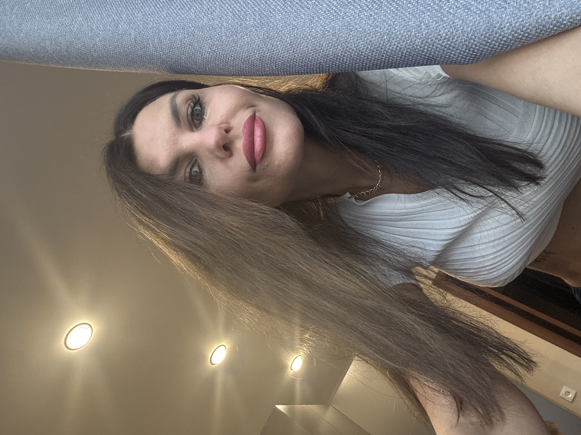 In a dimly lit room with recessed ceiling lights, a woman with long, dark brown hair and light green eyes poses for a selfie. She is wearing a white ribbed top and a gold chain necklace. Her makeup includes pink lipstick and visible eyelashes. To her right, a textured grey fabric surface is prominent. The background shows a wall with wooden paneling and a light switch. The image does not provide any visual information to determine the specific location within the city of Chaikovsky, Russia.