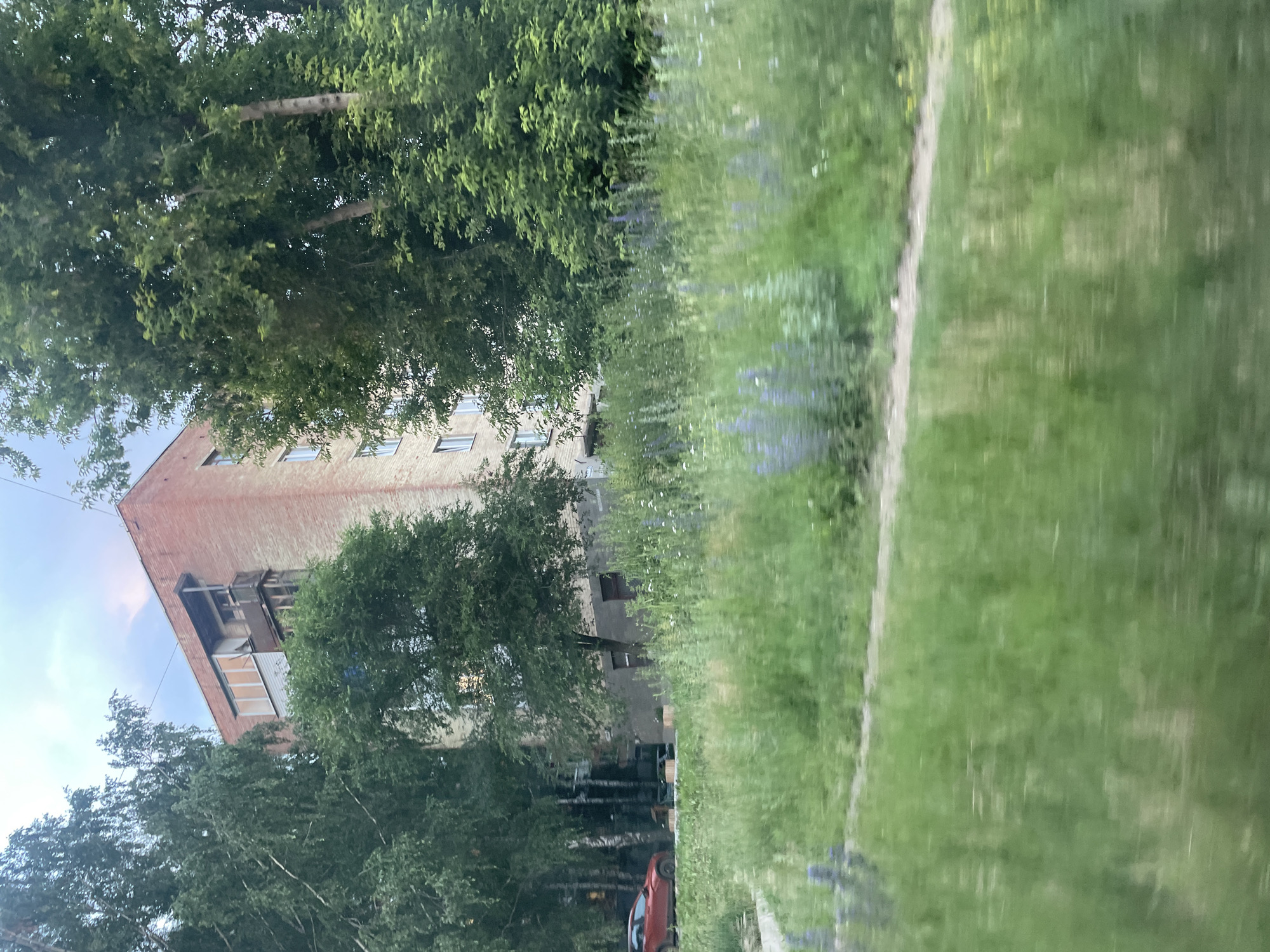 This media file captures a scene of an urban environment in Votkinsk, Russia. The primary subjects are a brick apartment building and surrounding trees and grass. The building, with its visible windows and balconies, appears to be multi-story and of a style common in Russian urban areas. Lush green trees partially obscure the building, with their leaves filling a significant portion of the frame. A patch of green grass with a visible path running through it occupies the right side of the image, suggesting a residential area or park. A red car is visible in the lower portion of the frame, parked near the building.
The image appears to be taken during the daytime, with the sky visible in the upper left corner indicating it's not nighttime. The lighting suggests it might be overcast or late afternoon/early morning, as there are no strong shadows. The overall mood is neutral, depicting a typical urban landscape. There is no discernible activity taking place beyond the parked car, and no people are visible. No text is present in the image. The orientation of the image is vertical.