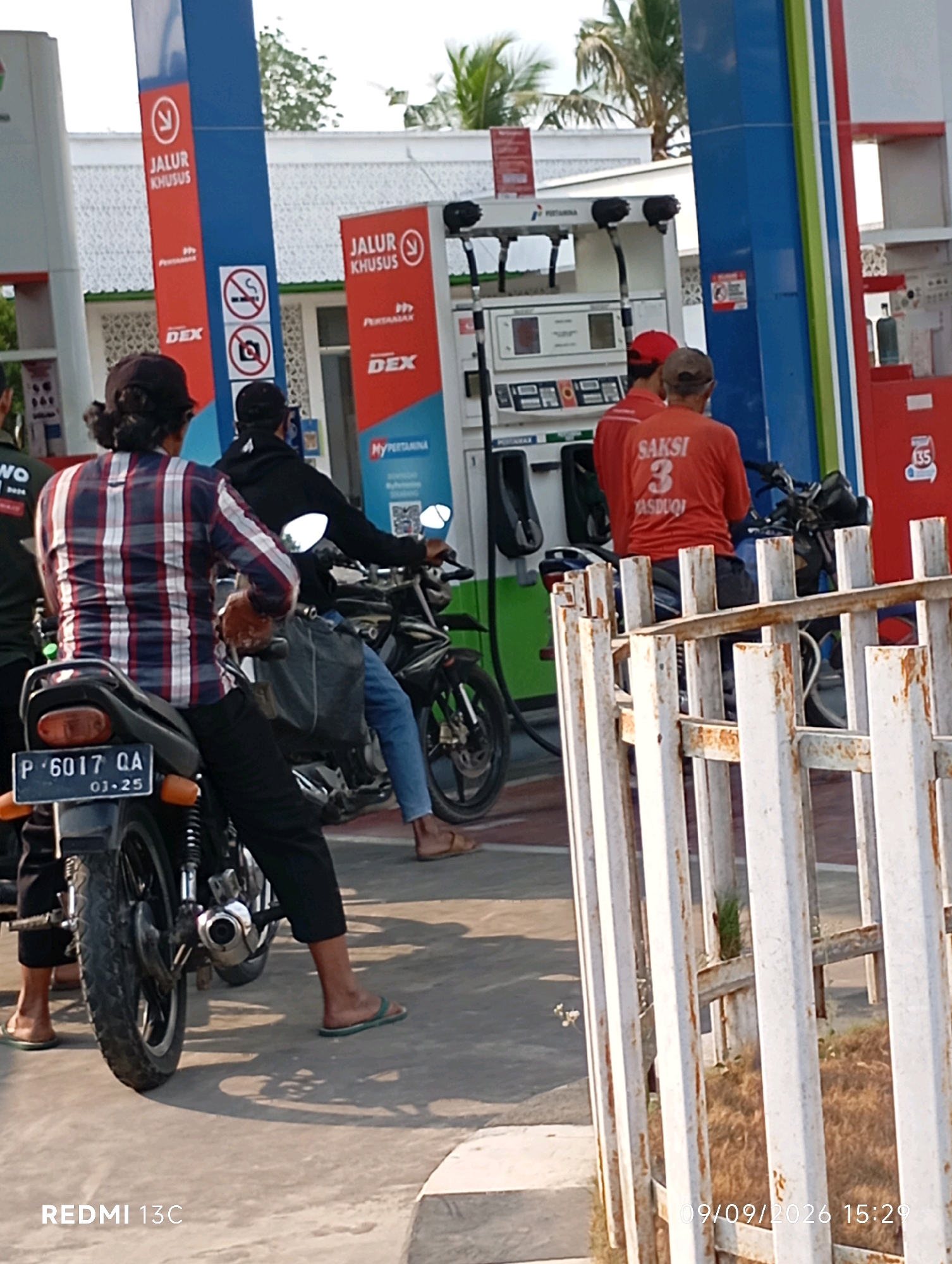 The image depicts a scene at a gas station in Rowotamtu, Indonesia. In the foreground, two individuals are seated on motorcycles. The person on the left is wearing a red, white, and blue plaid shirt and black pants. The person on the right is wearing a black hooded jacket and blue jeans. Both are facing away from the camera. Behind them, at a gas pump, two individuals in red shirts are visible. One is wearing a red cap, and the other is wearing a dark cap. One of the individuals in red has "SAKSI 3 YASDUQI" printed on the back of their shirt. The gas station features prominent blue and red signage with white lettering indicating "JALUR KHUSUS" (Special Lane) and "DEX" (Diesel Exhaust Fluid). There are also "no smoking" and "no photography" symbols visible. The environment appears to be outdoors, with a paved surface and a white metal fence in the right foreground. The lighting suggests it is daytime. A license plate on the motorcycle on the left reads "P 6017 QA 03-25". A timestamp at the bottom right of the image indicates "09/09/2026 15:29".