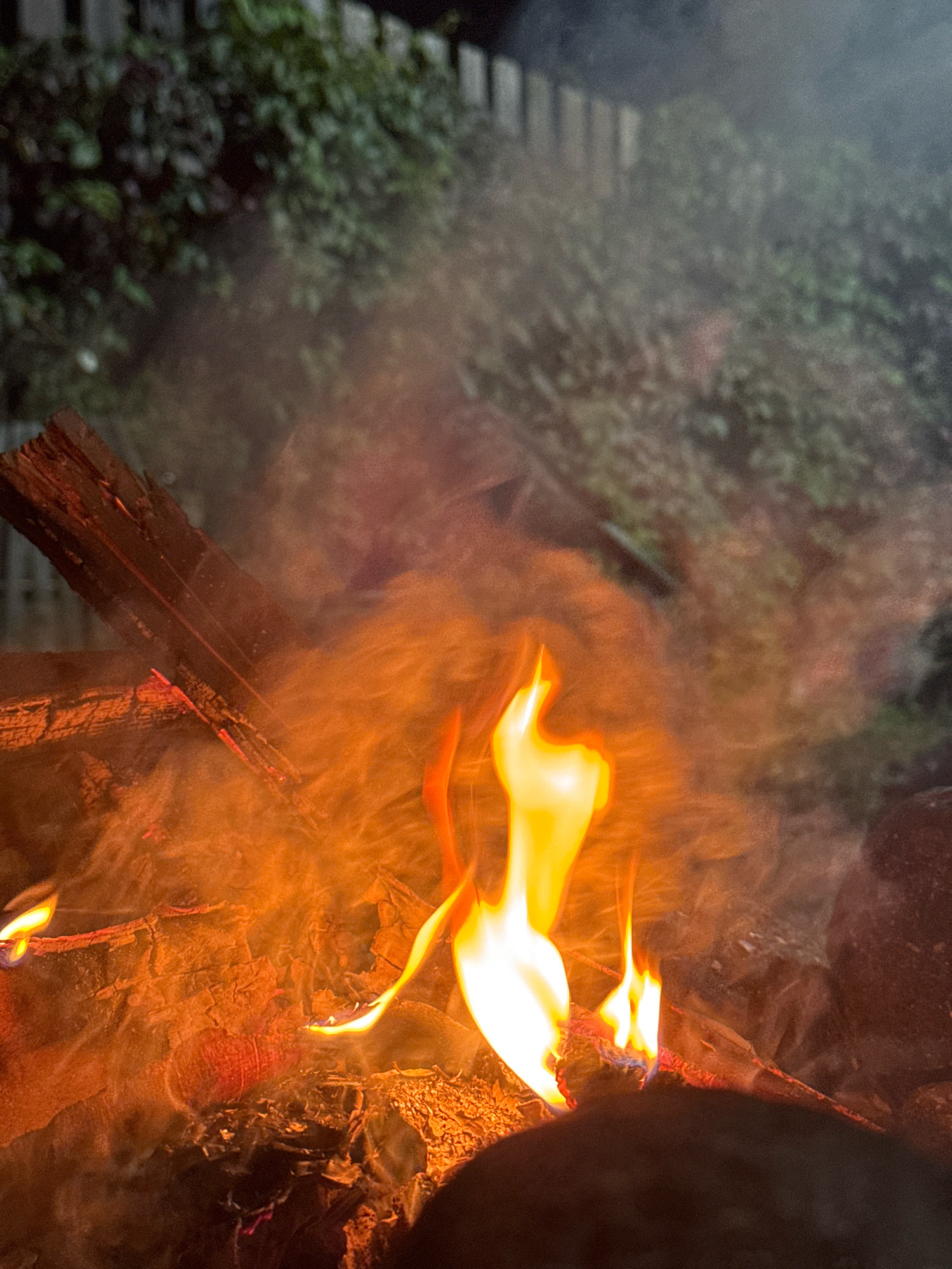 The image depicts a close-up view of a bonfire at night, likely in a backyard setting in Kempelevo, Russia. The fire is actively burning, with bright orange and yellow flames licking upwards. Smoke billows around the flames, partially obscuring the background. Several pieces of burnt wood are visible, some glowing with embers. In the background, a dark fence and lush green foliage can be seen, suggesting a natural or garden environment. The overall impression is one of a warm and lively gathering, with the fire serving as the central focus.
