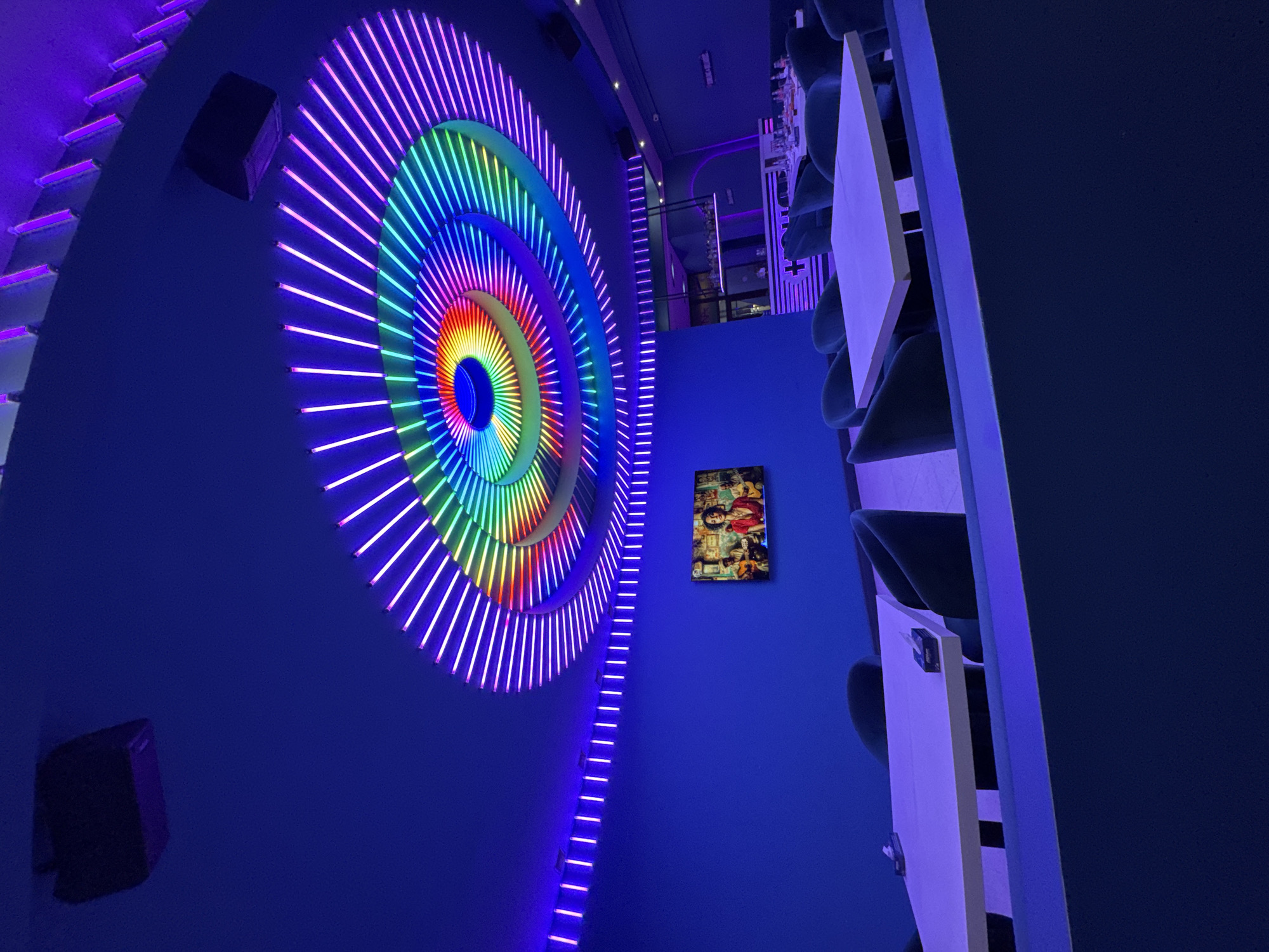 The image shows an interior space with distinctive lighting. A large, circular light display composed of concentric rings of neon lights in rainbow colors is mounted on a blue wall. To the right of this display, a rectangular picture frame hangs on the wall. Tables with white tops and chairs are arranged on the right side of the frame, oriented at an angle. A narrow strip of neon lights descends vertically along the wall, extending from the larger circular display towards the bottom. A black speaker is mounted on the upper left and another on the lower left. The image is shot from a low angle, exaggerating the height of the scene. The location is a restaurant in Yerevan, Armenia.