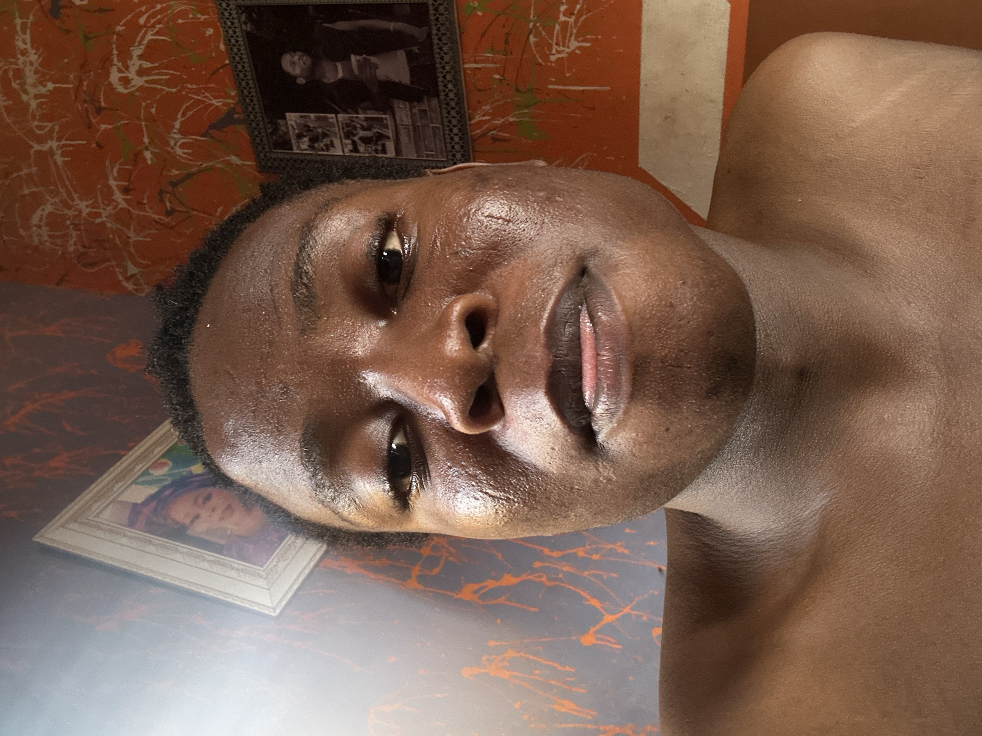 A close-up, tilted selfie of a dark-skinned young man with short, dark hair. He is shirtless. Behind him is a wall with orange and white paint splatters. There are two framed pictures on the wall: one in black and white above, and a color portrait lower left. The location is Unknown, Unknown.