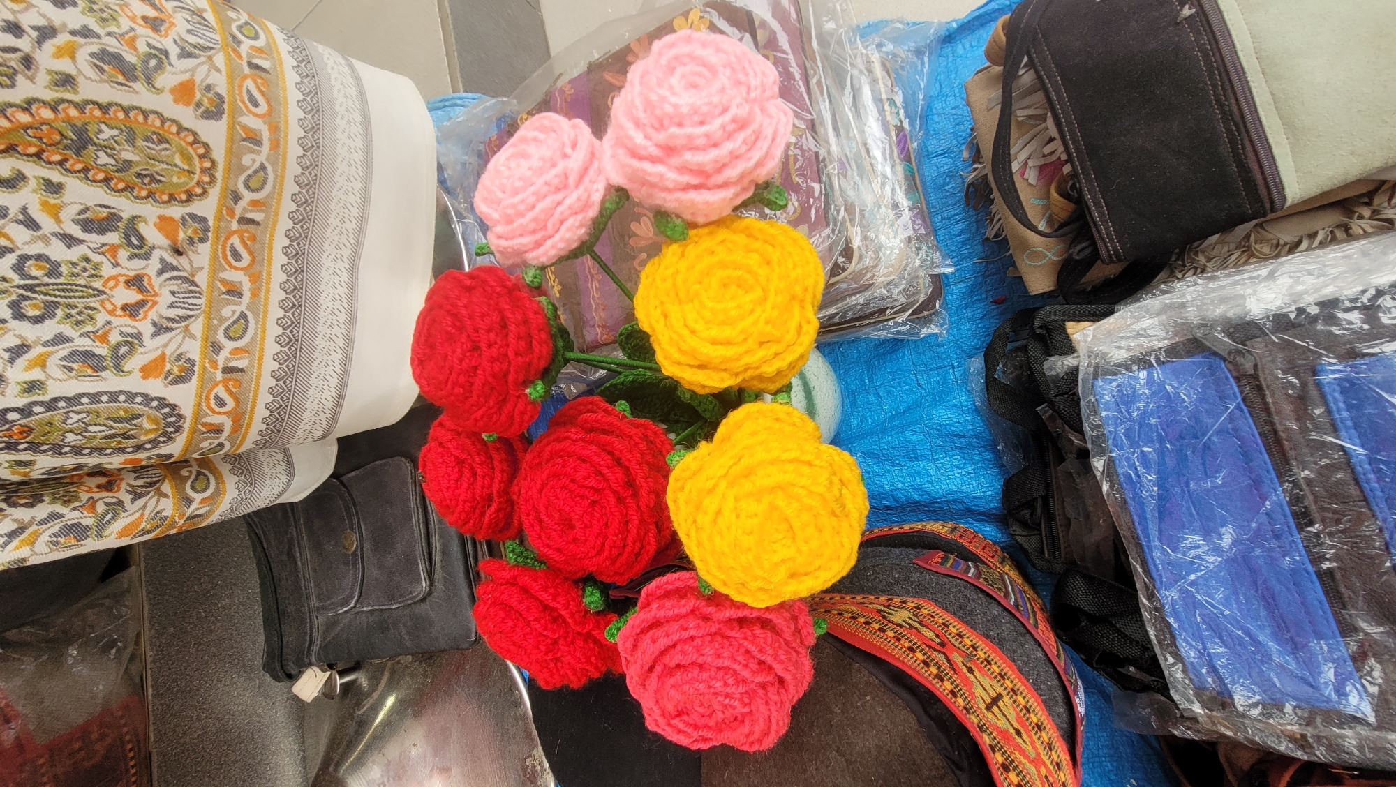 In Vashisht, India, a collection of hand-knitted woolen flowers in red, yellow, and pink hues are arranged in a white pot. These flowers are placed on a blue tarp, with various bags and textiles surrounding them. To the left, a black leather satchel with a front flap pocket is visible, as well as a dark-colored hat with a decorative woven band. Behind the flowers, a large textile with a white base and colorful paisley-like patterns is draped. To the right of the flowers, more textiles and bags are stacked, some enclosed in plastic packaging. The scene appears to be an outdoor market stall or a display of handicrafts.