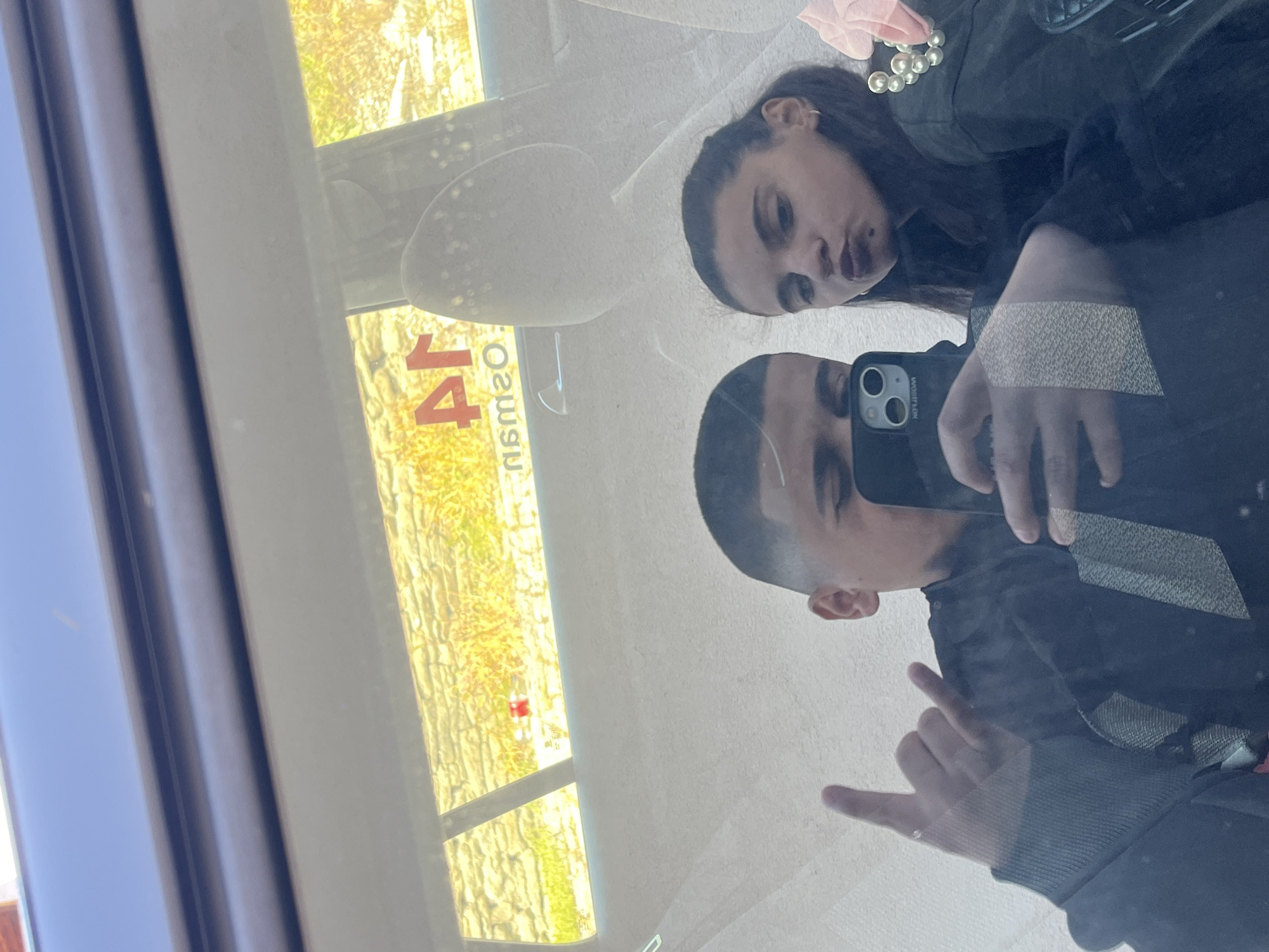 A selfie photograph is taken inside a vehicle. Two individuals are visible in the reflection of a window. The person on the left is a male with a shaved head, holding an iPhone in one hand and making a hand gesture in the other. The person on the right is a female with long, dark hair. They are positioned close together. Through the window, a street view is visible, and a sign with the number "14" is recognizable. The reflection suggests the vehicle is stopped. The location is Lozarevo, Bulgaria.