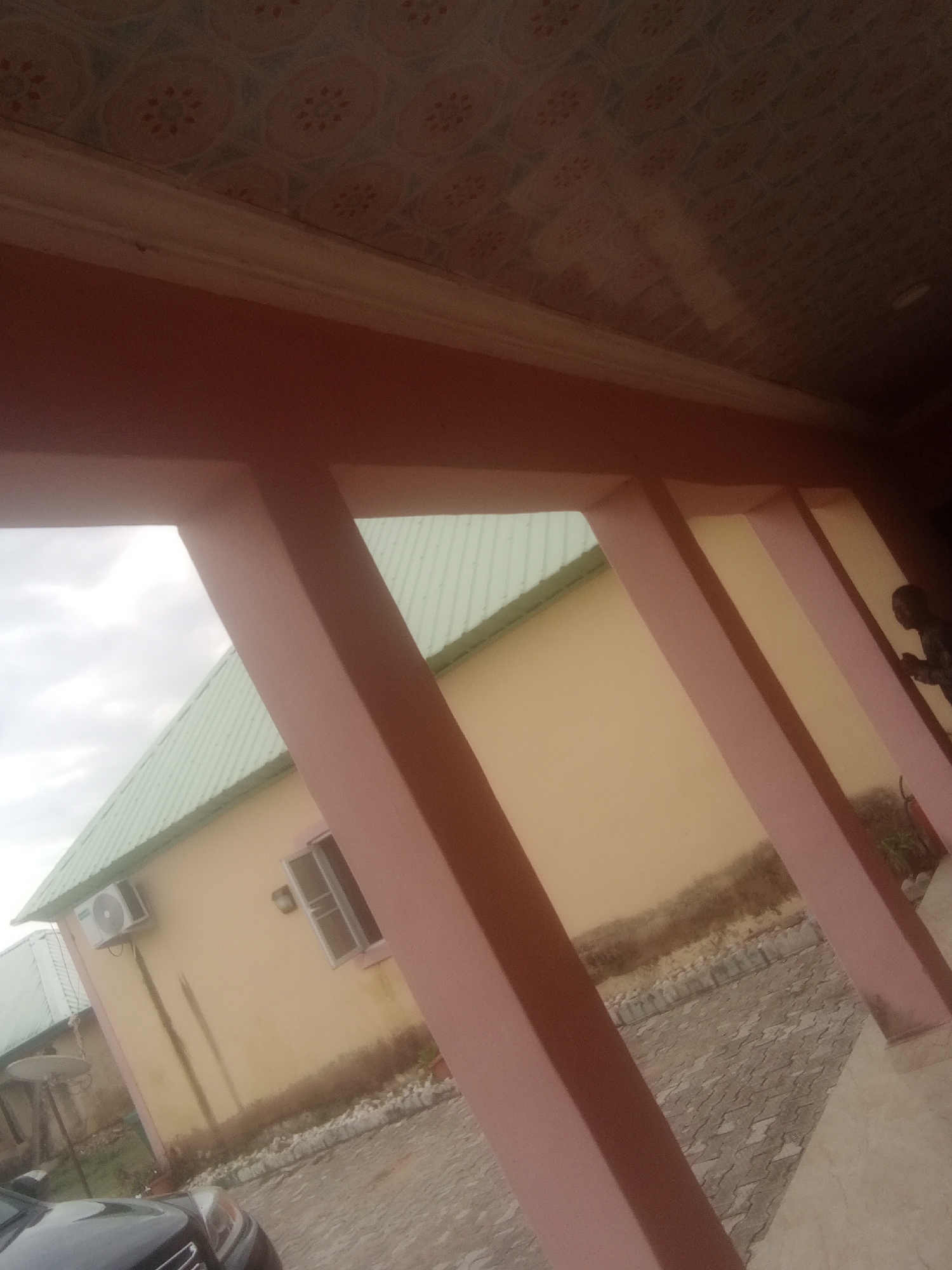 The image shows a building with a light yellow facade and a green corrugated metal roof. A light pink covered porch with several support beams runs along the front of the building. An air conditioning unit is mounted on the exterior wall to the left of a window. The ground in front of the building is paved with interlocking stones. In the distance, a portion of a dark-colored car is visible. A person in silhouette is visible to the far right, with their hands clasped in front of them. The sky is overcast, suggesting it is either early morning or late afternoon. The scene appears to be residential or commercial property. There is no visible text in the image. The location context is Jalingo, Nigeria.