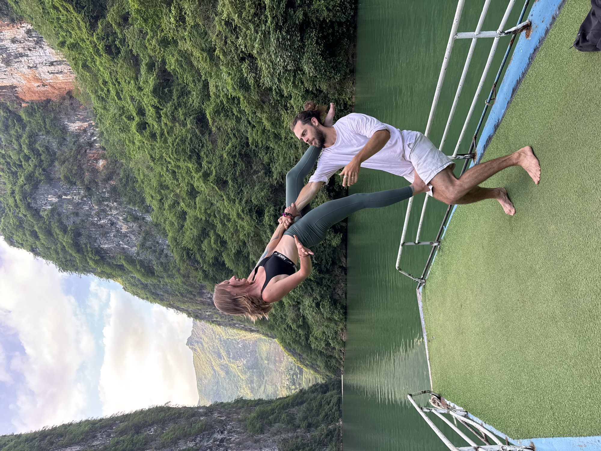 Two people are performing acro yoga on a boat, with a green river and steep, vegetated cliffs as the backdrop.
A man with a beard and hair in a bun wears a white t-shirt and white shorts. He's standing on the boat's green carpet, barefoot, holding the hands of a woman and supporting her.
A woman wears a black sports bra and green leggings. She is upside down, her legs extended upward, held by the man.
The boat has a blue and white railing. The river is a greenish color, and the cliffs are covered in lush vegetation. The sky is visible with clouds in the background.