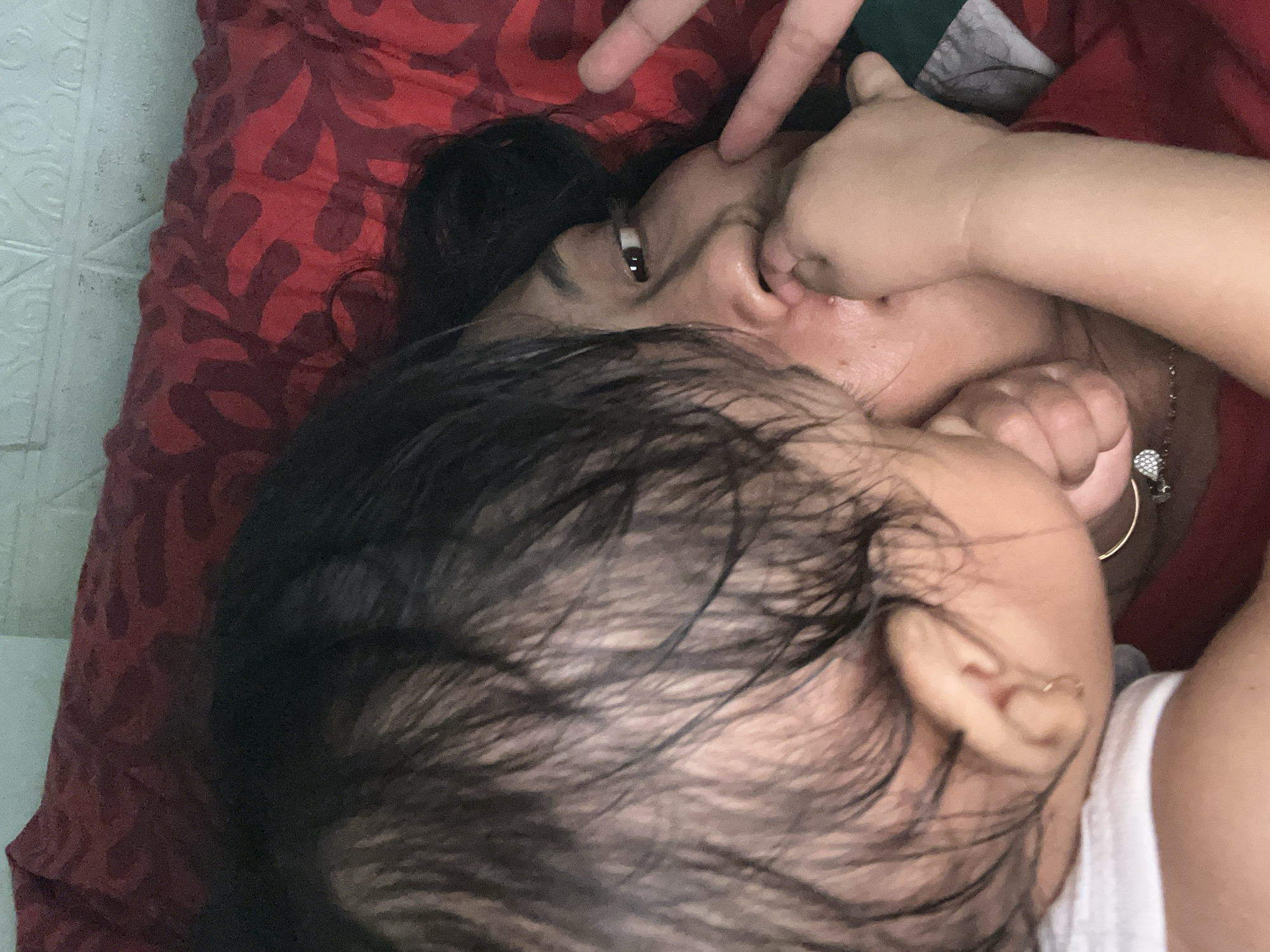 A woman lies on a red patterned cushion, her face partially obscured by the head of a small child with dark hair. One of the woman's eyes is visible, and she appears to be interacting with the child. The location is Kecamatan Pati, Indonesia.