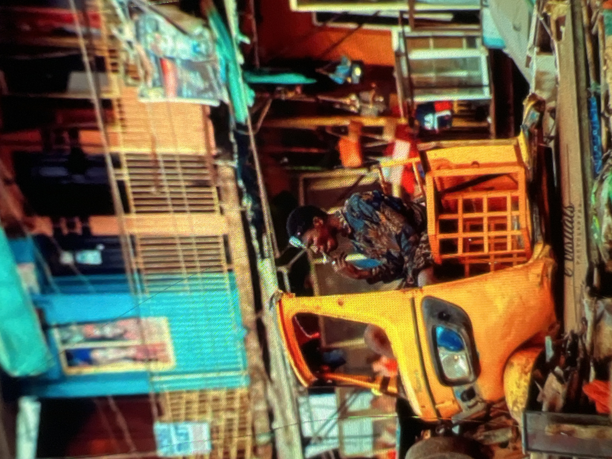 In a bustling urban scene in Lagos, Nigeria, a man is seated in the driver's seat of a bright yellow auto-rickshaw, commonly known as a "Keke Napep." He is holding a cigarette to his mouth and appears to be smoking, with his head tilted slightly as he looks off to the side. The man is wearing a dark, patterned long-sleeved shirt and a headpiece. The Keke Napep has a prominent headlight and a metal grille or cage at the rear. Behind the auto-rickshaw, the environment is crowded and appears to be a market or commercial area with buildings constructed from various materials, including corrugated metal and painted facades in vibrant colors like teal and orange. The buildings have multiple levels, with windows and balconies visible. Various objects and items are stacked and displayed, suggesting commercial activity. The overall scene depicts the dynamic and lively atmosphere characteristic of Lagos street life.