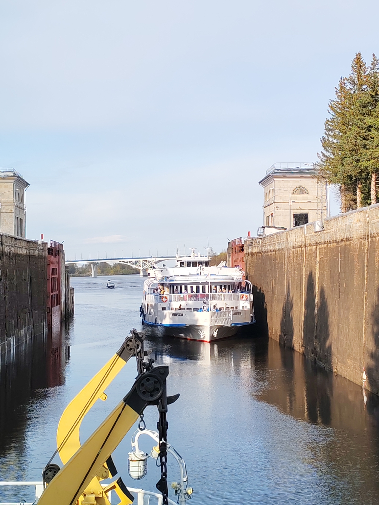 The image shows a large white cruise ship navigating through a lock system. The lock walls are made of gray concrete and flank the ship on either side. In the foreground, part of a yellow crane is visible on the deck of another vessel, likely the one from which the photo was taken. In the background, a bridge spans across the water, with another smaller boat visible in the distance. The sky is light blue with some clouds. The setting appears to be an urban waterway.