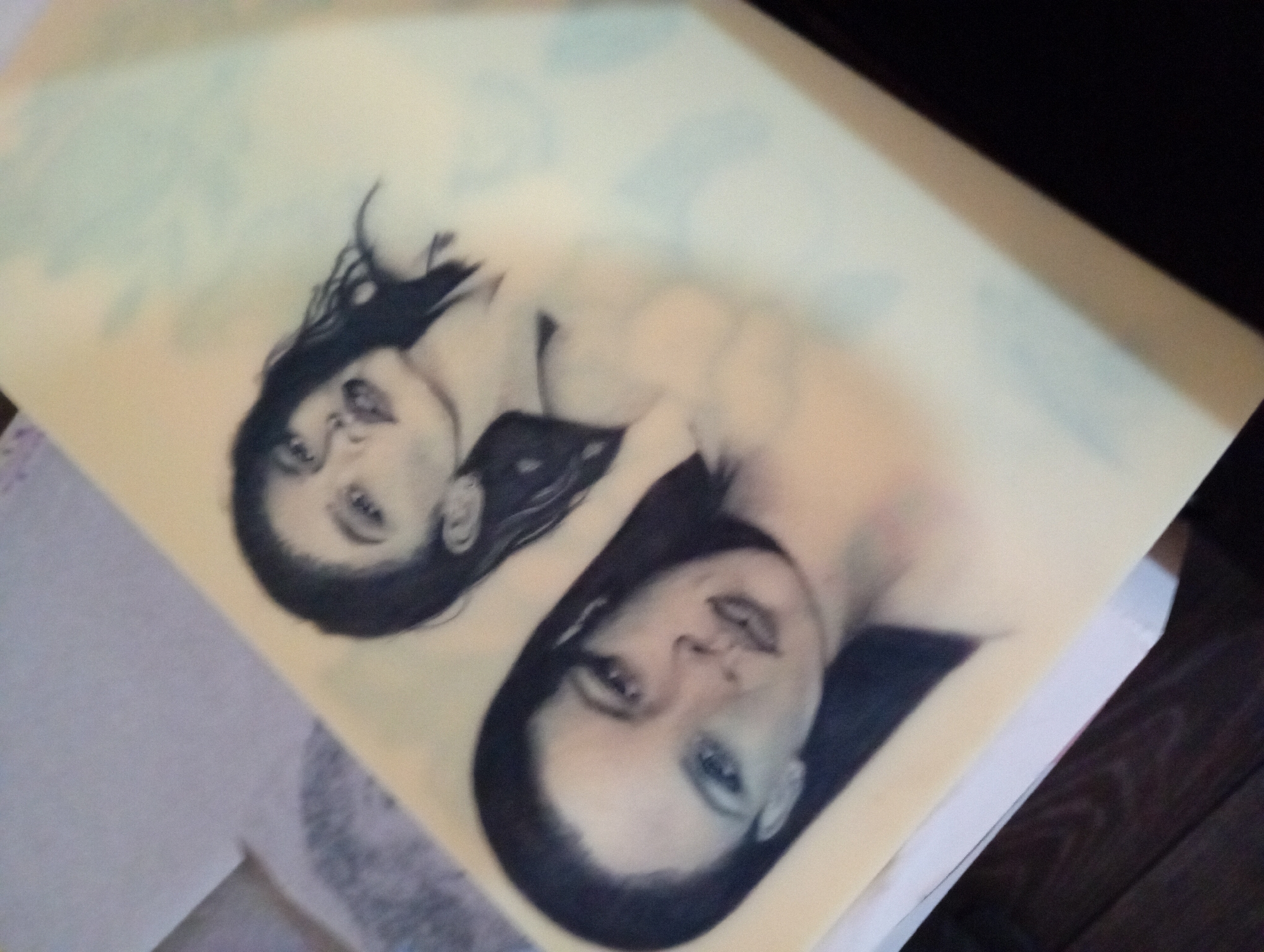 The image depicts two portraits of women printed on a large sheet of paper. The portraits are rendered in grayscale, with dark hair and lighter skin tones. The women have similar facial features, including prominent eyes and lips. The sheet of paper is light yellow or beige, and the portraits are oriented diagonally. The background is partially visible, including a wooden floor and a white surface, suggesting the image was taken indoors. The location in the city of Roza, Bulgaria, is not discernible from the image.