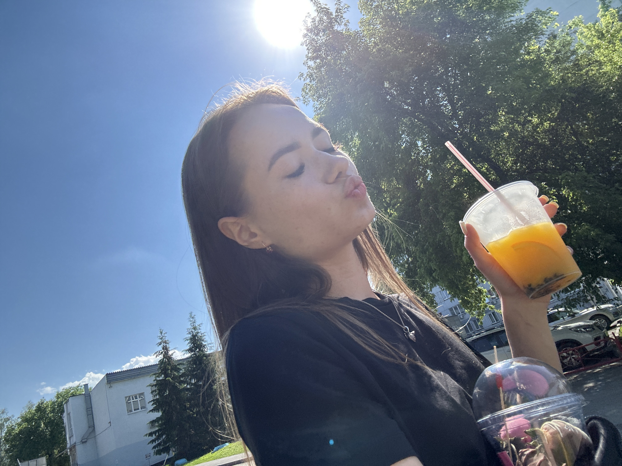 A young woman with long brown hair is captured in a close-up, outdoor selfie. She is leaning her head back slightly, with her eyes closed and lips pursed as if to blow a kiss. She is holding a clear plastic cup filled with an orange beverage and ice, with a pink straw. The background features a bright blue sky with a sunbeam shining down, a large green tree, and a building with windows. Another clear plastic cup with a pink lid and what appears to be a straw is visible in the foreground. The woman is wearing a black t-shirt and a silver necklace. The lighting suggests it is daytime and sunny.