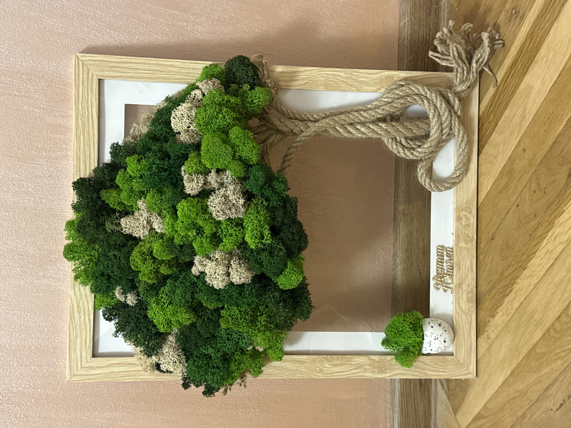 The image shows a moss-covered decoration hanging on a wall. The decoration is framed by a light wood frame with a white inner border. The moss is predominantly green with patches of cream color, resembling foliage. A thick rope is looped around the upper right corner of the frame, its ends frayed. A small pot containing moss sits in the lower right corner of the frame. A small engraved sign with the word "Harmony" written in a Cyrillic alphabet font is located on the lower right side of the white border. The background walls are textured and have different colors, one is peach colored and the other is wood grained. The location is implied to be in Plovdiv, Bulgaria, based on the Cyrillic text on the sign. There are no people visible in the image.