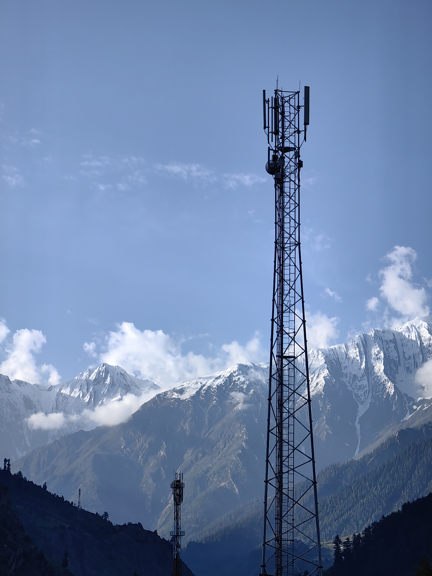 A tall, lattice telecommunications tower stands in silhouette against a bright blue sky with scattered white clouds. The tower is positioned on the right side of the frame and features multiple antennas mounted at its apex. In the background, snow-capped mountains rise, partially obscured by lower, darker mountain slopes covered in trees. Another, smaller telecommunications tower is visible in the distance on the left, situated on a darkened hillside. The scene is a landscape view, likely in a mountainous region. The location is not Udaipur, India.