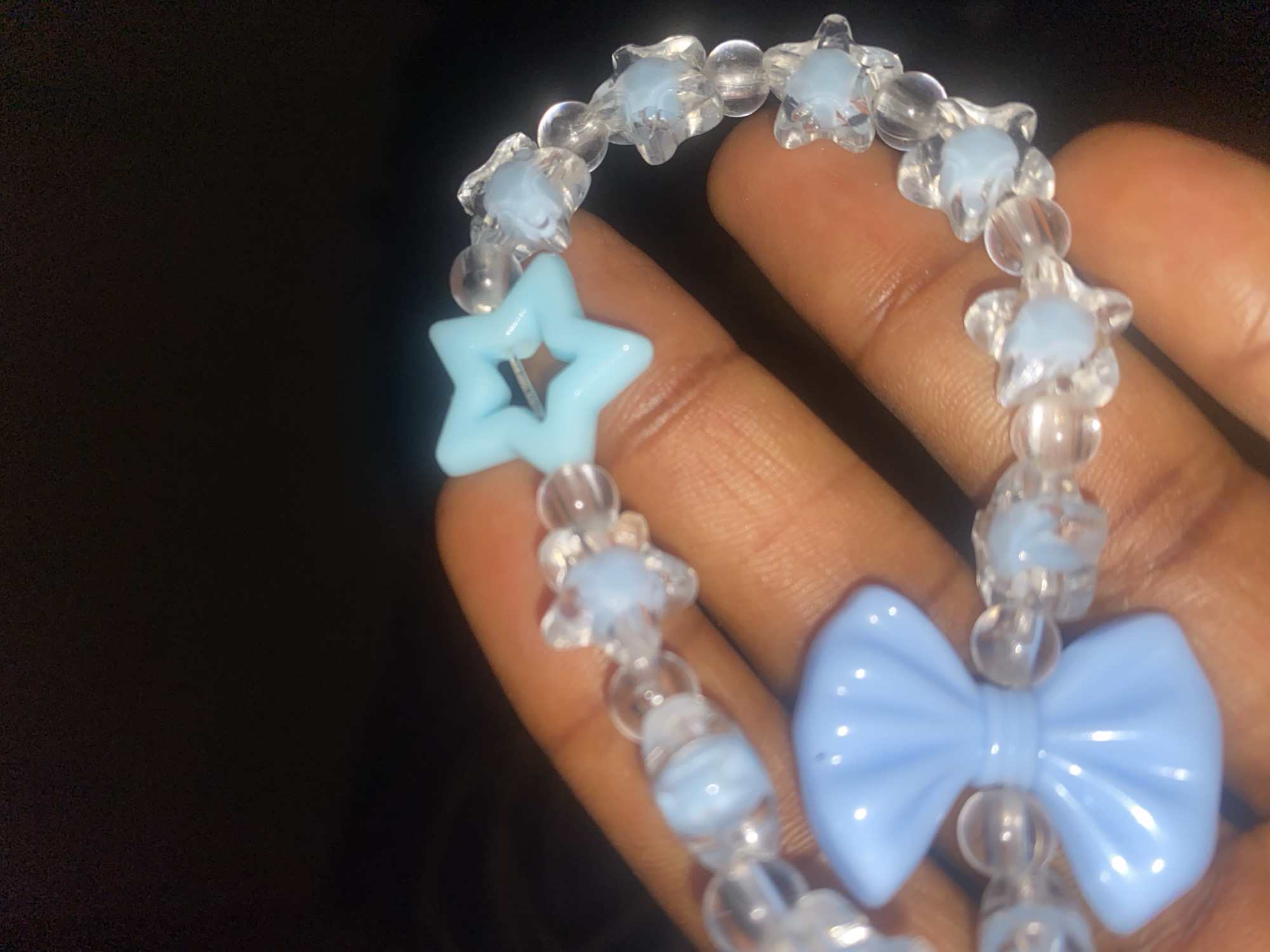 The image displays a close-up of a person's hand holding a light blue and clear beaded bracelet. The hand, with brown skin tone, occupies the right side of the frame, providing a dark background for the bracelet.
The bracelet features a repeating pattern of clear round beads interspersed with clear, star-shaped beads that have a soft, light blue center. A larger, hollow, light blue star-shaped bead is prominent on the left side of the bracelet, through which the elastic string is visible. Further down, towards the bottom right, a distinct light blue bow-shaped bead is visible, serving as a focal point.
The lighting is subdued, creating a dark, out-of-focus background, which highlights the shiny, translucent quality of the beads. There is no discernible activity other than the hand presenting the bracelet. No text is visible in the image. The scene is likely set indoors in Jos, Nigeria, given the provided location context.