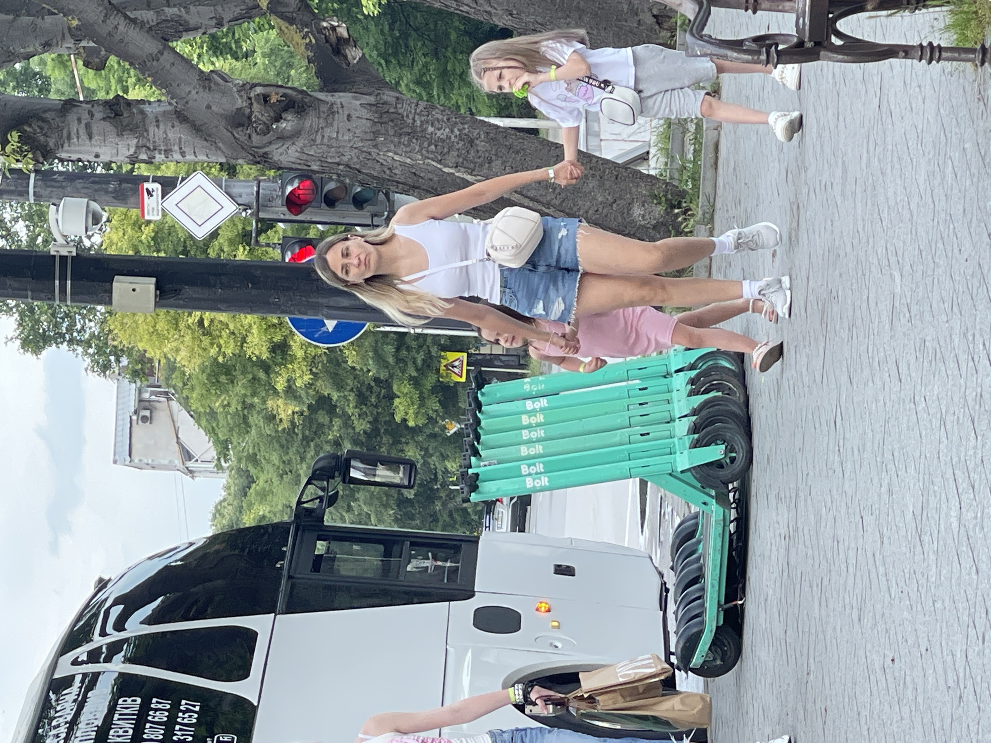The image depicts an urban pedestrian scene in daylight. In the foreground, a blonde adult woman wearing a white tank top, distressed denim shorts, white sneakers, and a white crossbody bag walks forward, holding hands with two young girls. She also wears a neon yellow wristband on her left wrist.
To her left, a young girl in a pink dress and sandals is partially visible, holding the woman's left hand. She also wears a neon yellow wristband. To the adult woman's right, a young girl with light brown hair, wearing a white t-shirt with a "Pusheen" cat graphic, grey shorts, and white sneakers, holds the woman's right hand. This girl also has a white crossbody bag and a neon yellow wristband on her left wrist, and she holds a small, green object to her mouth.
In the mid-ground, immediately behind the adult woman, a row of approximately eight bright green electric scooters, each marked with "Bolt" in white lettering, are parked perpendicular to the direction of travel. To the left of these scooters, the rear and side of a large white and black passenger bus are visible. Text in Cyrillic, including "КВИТКІВ" (tickets) and phone numbers "807 66 87" and "317 65 27", is visible on the bus's side. A partially visible individual wearing a white tank top stands near the bus, holding two brown paper shopping bags, one of which displays the "ZARA" logo.
To the right of the walking group, a thick-trunked tree with gnarled bark stands. Further right, a partial view of a dark metal bench is visible. In the background, behind the people and scooters, a black utility pole supports a white security camera, a red-bordered warning sign with a camera icon, a white diamond-shaped traffic sign (indicating priority road), and a traffic light showing a red signal in both upper lenses. A circular blue traffic sign is also visible below the warning sign.
The ground is paved with grey interlocking pavers. Beyond the immediate street infrastructure, green trees and foliage are visible, along with parts of distant buildings under a partly cloudy sky. The overall impression is an active urban environment.