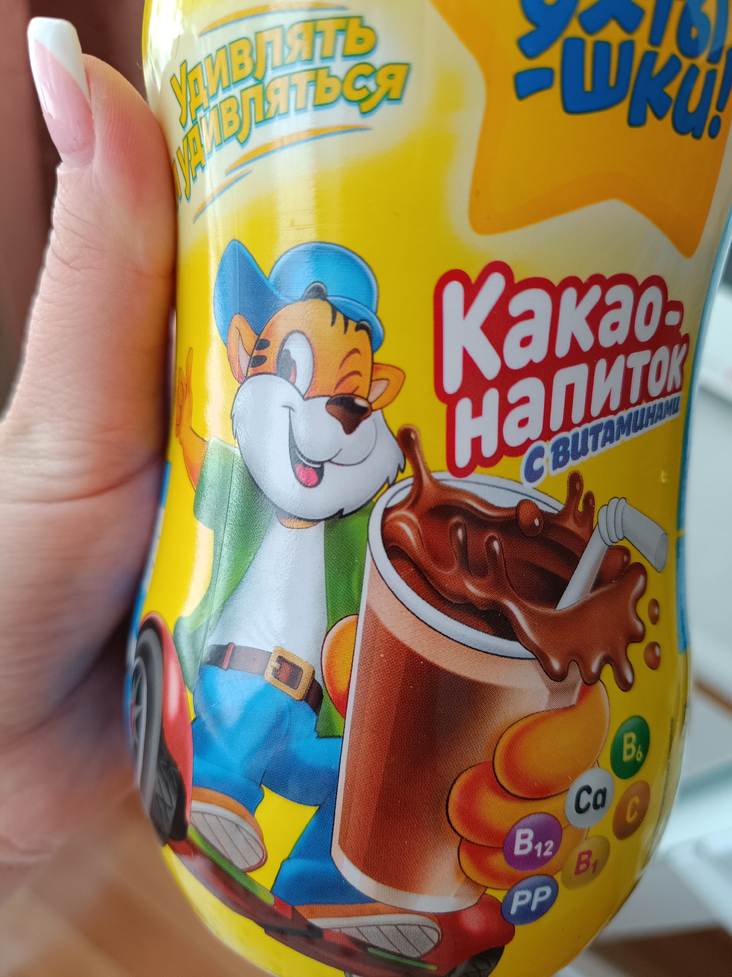 A hand is holding a bright yellow container featuring a cartoon tiger character on it. The container has Russian text, including the words "Какао-Напиток С Витаминами" (Cocoa Drink with Vitamins). The container also displays representations of vitamins, such as B6, Ca, C, B12, B1, and PP. The overall design is playful and aimed at children. There is no evidence of Tobolsk, Russia.