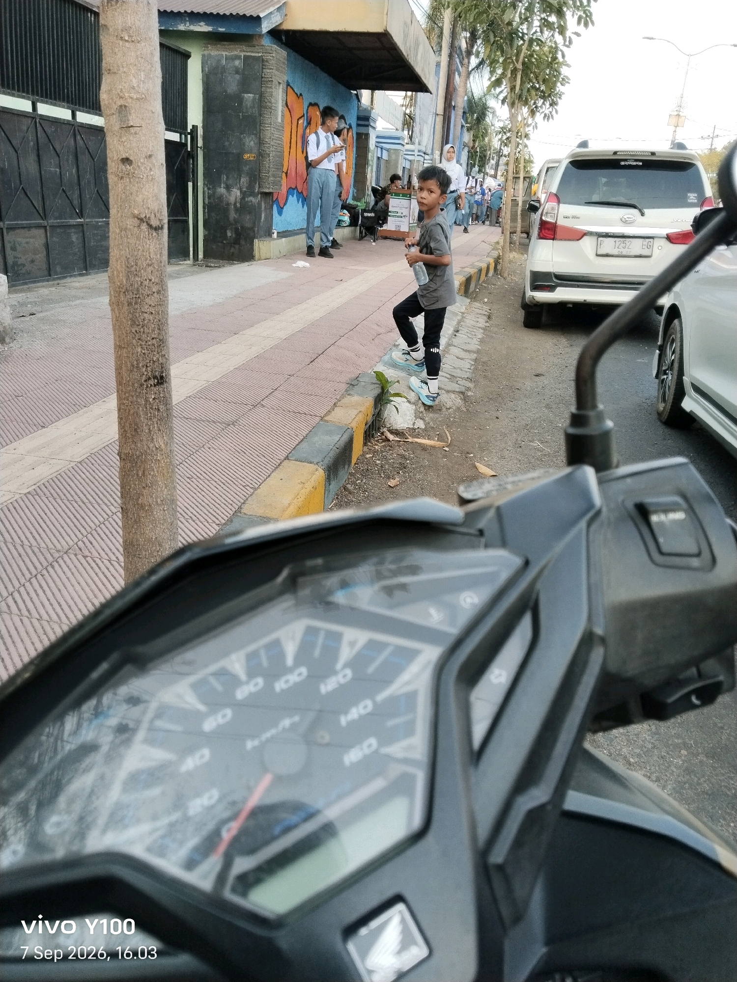 The image depicts a street scene in Situbondo, Indonesia, likely taken from the perspective of a parked motorcycle. In the foreground, a portion of the motorcycle's dashboard is visible, showing a speedometer with a needle pointing slightly above 20 km/h, along with other indicators. The date stamp "7 Sep 2026, 16.03" suggests the time and date the photo was taken.
To the left, a tree trunk stands prominently, partially obscuring a black metal gate and a building with a blue and white facade. A vibrant mural adorns the wall of the building. Further down the sidewalk, two students in school uniforms, one holding a phone, are standing near the mural.
A young boy, dressed in a gray t-shirt and black pants, stands on the sidewalk, holding a bottle of water and looking towards the camera. Behind him, several people are walking along the sidewalk, and a white SUV is parked on the street. The environment appears to be a public street with a sidewalk, curb, and paved road. The sky is bright, indicating daylight.