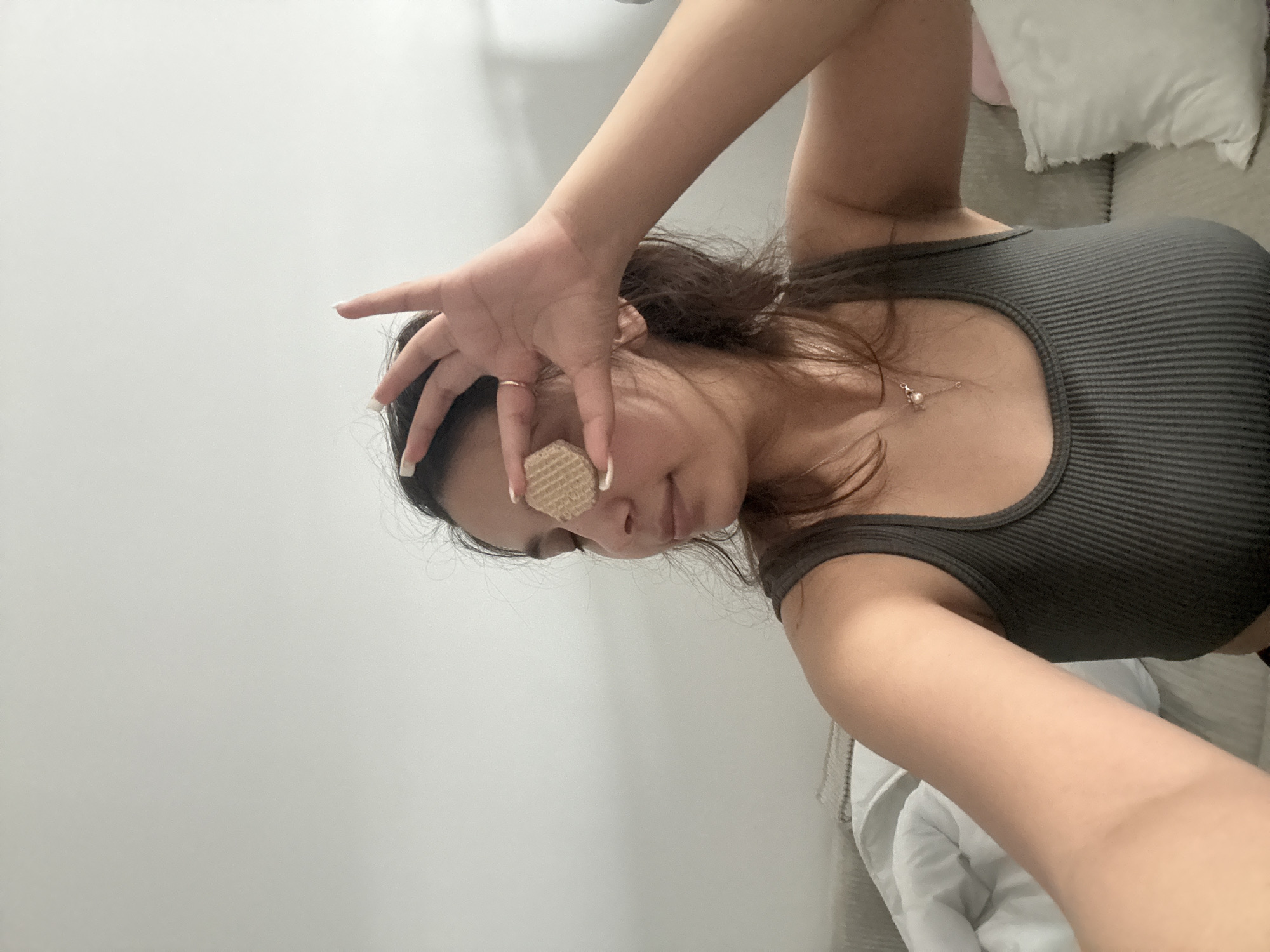 A woman is holding a round, beige wafer-like object over one eye, obscuring it from view. She has dark hair pulled back and is wearing a grey ribbed tank top and a delicate necklace. Her skin appears light-toned, and she has white-tipped manicured nails. The background includes a plain white wall and, in the periphery, a white pillow or bedding on a grey upholstered surface. The image appears to have been taken indoors. The location cannot be determined from the provided image.