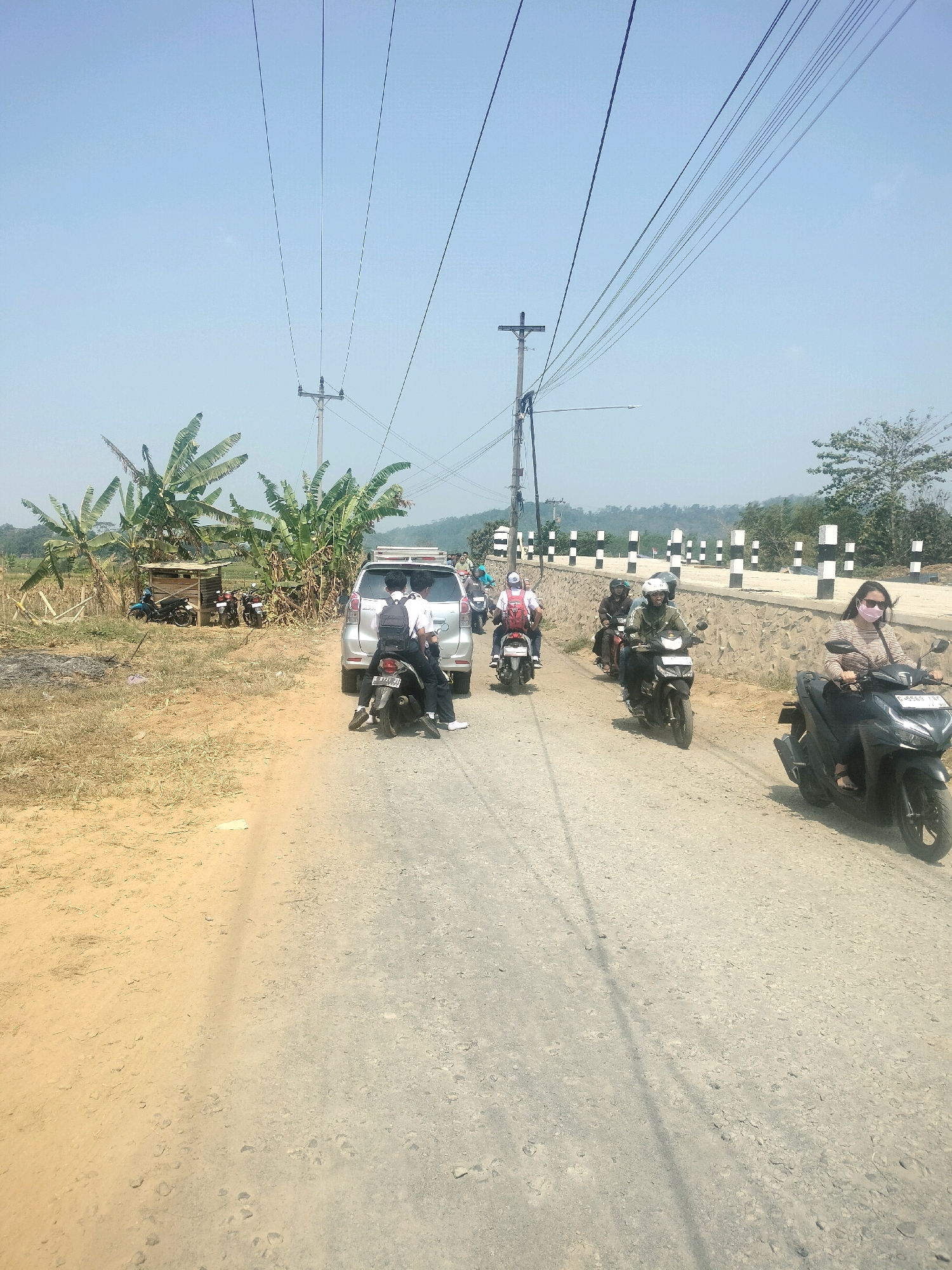 The image depicts a rural road scene in Tersono, Indonesia, under a clear blue sky. A convoy of motorcycles and a silver car are traveling on a dusty, unpaved road. In the foreground, a woman wearing sunglasses and a pink face mask rides a black motorcycle. Behind her, several other motorcycles follow, some carrying multiple passengers. Two young men on a motorcycle, one with a backpack, are in front of the silver car. To the left of the road, a small wooden structure is visible near banana trees and dry vegetation. Further down the road, a stone wall with alternating black and white posts lines the side. Overhead, power lines crisscross the sky, supported by utility poles. The overall environment suggests a dry, sunny day in a sparsely populated area.