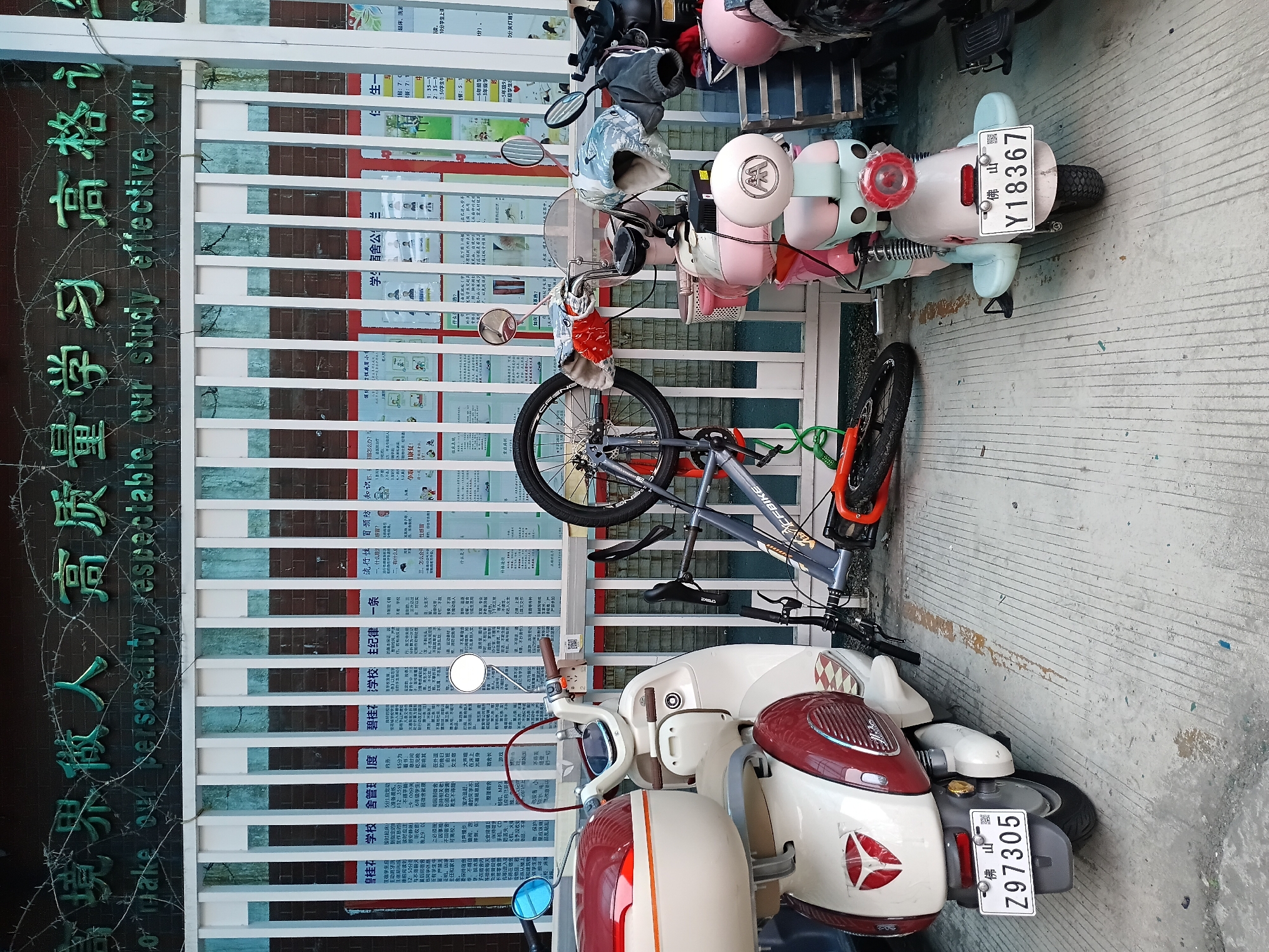 The image captures an outdoor scene, likely in front of a building, where multiple two-wheeled vehicles are parked. In the foreground, a white and maroon scooter is visible, bearing the license plate "佛山 Z97305". Above and behind it, a grey bicycle with orange accents is parked, featuring "CFBIKE" on its frame. Further back, a light blue and pink scooter with the license plate "佛山 Y18367" is present. Helmets and other personal items are seen placed on some of the scooters.
The background features a building wall that transitions from a dark surface with raised green Chinese characters to a white slatted fence. The green text on the dark wall reads "境界做人高质量学习 高格", with a partial English translation beneath: "To make our personality respectable, our study effective, our". The white fence is adorned with numerous Chinese posters, which appear to be school-related announcements or rules. Visible text snippets from these posters include "桂花 学校舍管度" (Guihua School Dormitory Management Regulations), "碧桂花学校主纪律一条" (Main Discipline Article of Guihua School), and "流行性冒预防知识匹、怎么办?" (Knowledge for Influenza Prevention: What to do?). The ground is a light-colored concrete pavement. The scene takes place during the daytime.