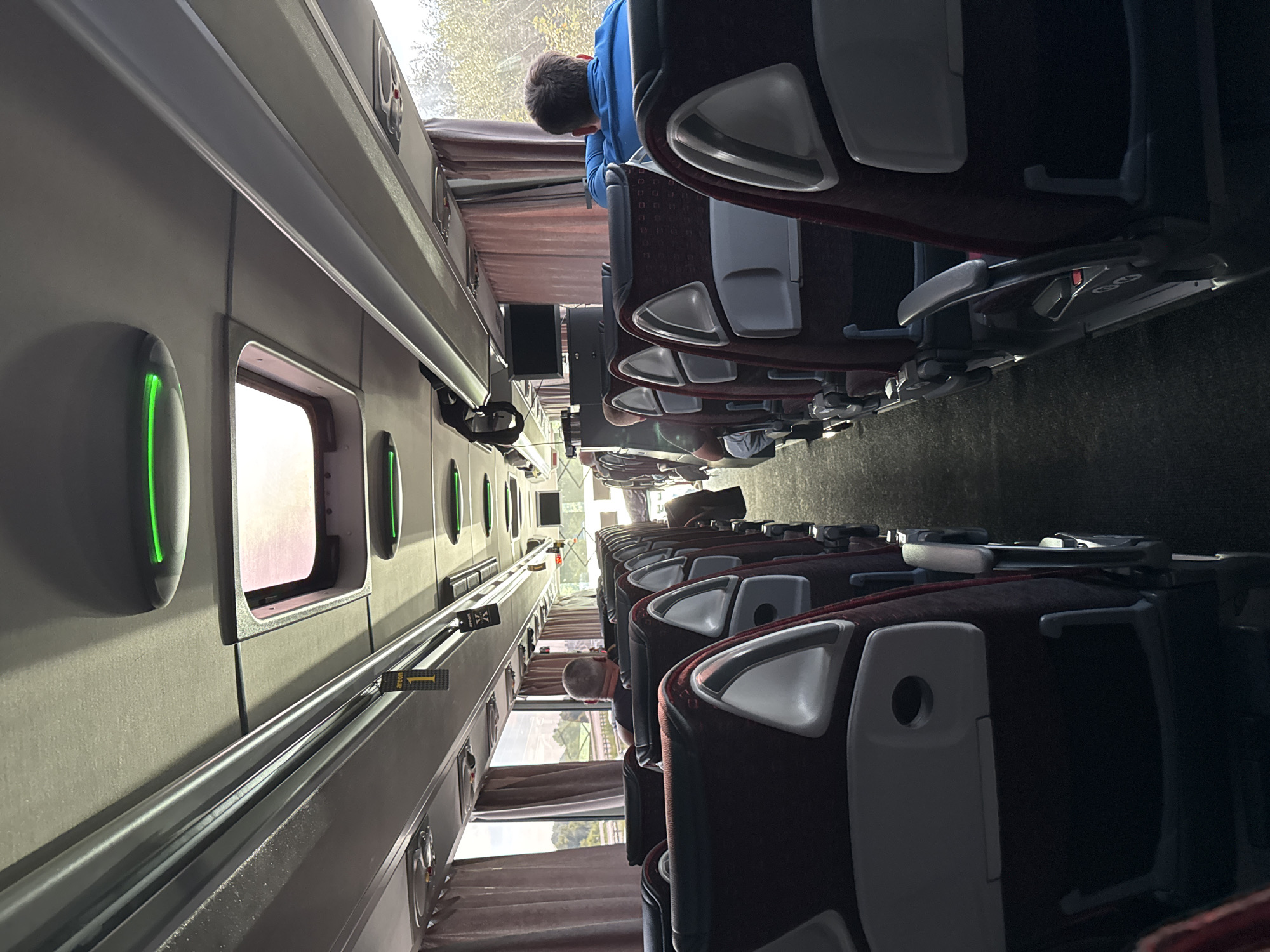 The image shows the interior of a bus with rows of empty seats. The seats are upholstered in a dark red fabric with light gray headrests and armrests. The aisle is carpeted in a dark gray material. The bus has large windows on either side, with light pink curtains drawn partially open. The ceiling of the bus features overhead storage compartments and individual reading lights, some of which are illuminated with a green glow. There are also skylights in the ceiling, allowing natural light to enter. A few passengers are visible in the seats, facing forward, including a man in a blue shirt on the right and a man in a dark shirt on the left. The overall lighting suggests it is daytime. A tag with the number "1" is hanging from an overhead rail. The environment appears to be a public transportation setting.