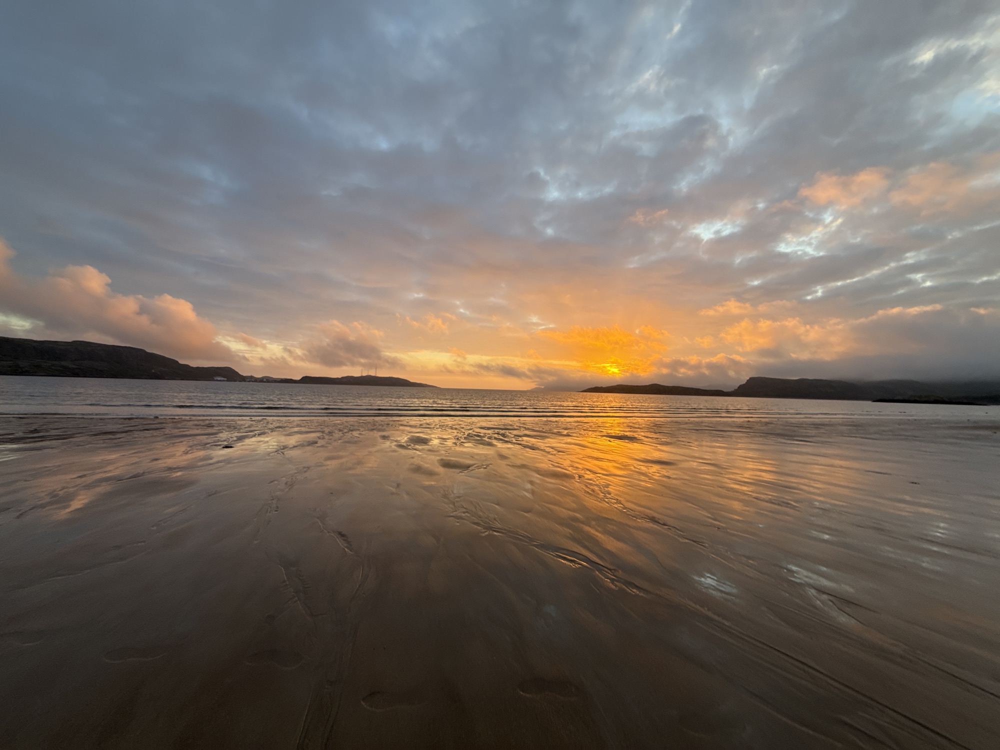 The image depicts a wide, sandy beach at sunset in Teriberka, Russia. The sky is filled with dramatic clouds, colored in shades of orange, yellow, pink, and gray. The setting sun casts a brilliant orange glow across the horizon, reflecting vividly on the wet sand and the calm ocean. The foreground is dominated by the wet sand, which shows patterns of ripples and textures left by receding waves. Distant landmasses, appearing as dark silhouettes, frame the scene on the left and right sides. The overall atmosphere is serene and visually striking, characteristic of a coastal sunset.