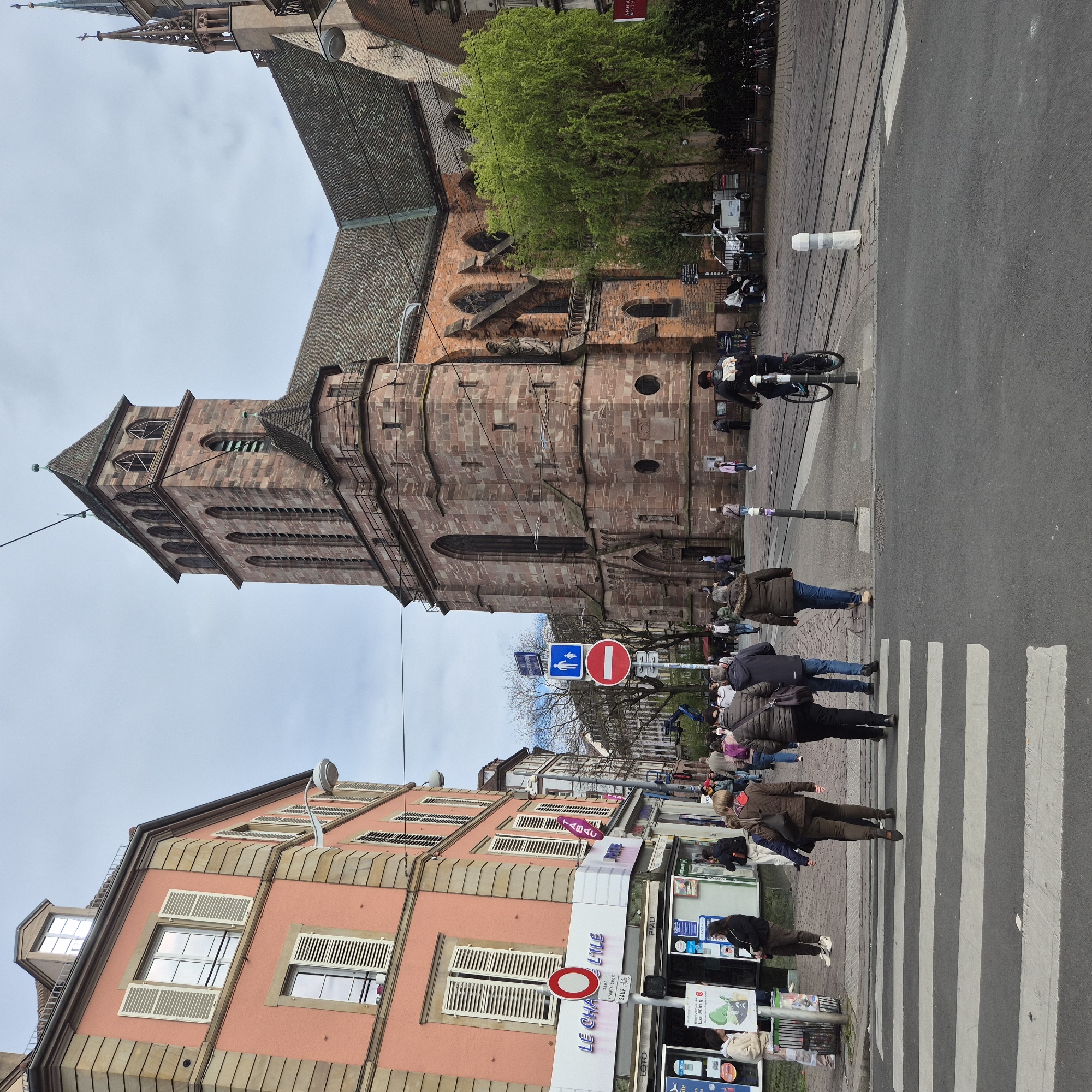 Here is a description of the image:
The image shows a street scene in Strasbourg, France, with a prominent view of the Église Saint-Pierre-le-Jeune. A pedestrian crosswalk dominates the foreground where several people are crossing. The church is on one side of the street, characterized by its red sandstone construction and tall tower. On the opposite side of the street, there are buildings with a mix of architectural styles, including shops and residential structures, one bearing the name "LE CHA L'ILE". Street signs and traffic control devices are visible. There are several people crossing the street.