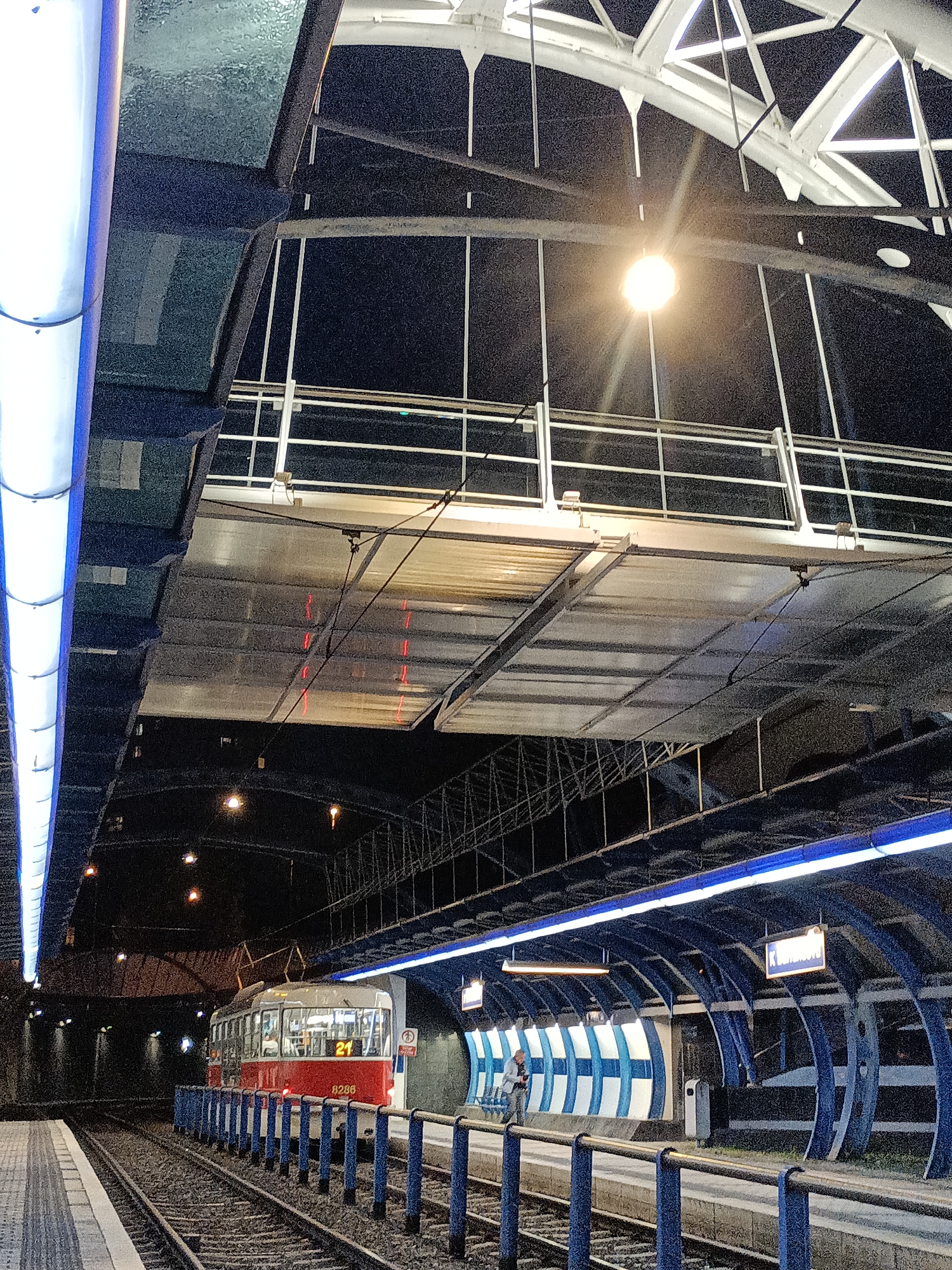 The image depicts a tram stop at night in Prague, Czechia. The scene is illuminated by artificial lights, including long, blue-lit panels running along the platform roof and a bright overhead spotlight. A red and white tram with the number "21" displayed on its front is stopped at the platform, with its headlights on. The tram is marked with the number "8286".
The tram stop features a modern, elevated design with a white, arched roof structure and a transparent canopy over the platform. Blue railings separate the platform from the tram tracks. In the background, the platform curves away, with a sign visible that reads "K Lihovaru". A single person is standing on the platform, looking towards the tram. The overall atmosphere is urban and functional, with a sense of quiet awaiting the tram's departure. The time of day is clearly night due to the darkness and artificial lighting. There are no visible weather cues, but the setting suggests an urban transport hub.