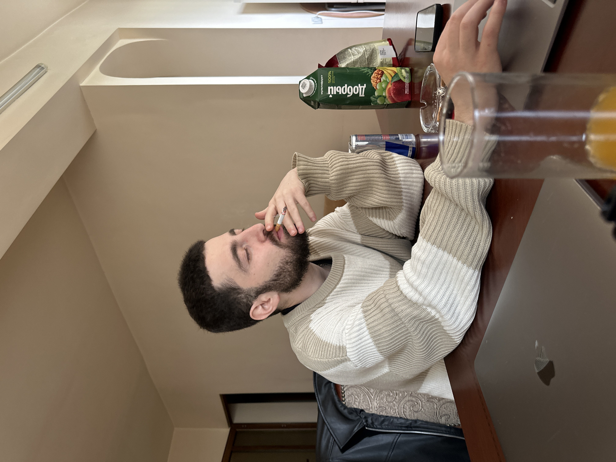 A young man with a beard and dark hair is depicted sitting at a wooden table in an indoor setting in Yerevan, Armenia. He wears a striped white and beige knitted sweater and is seen actively smoking a cigarette, with a faint wisp of smoke visible. His eyes are closed or cast downwards, suggesting a moment of contemplation or relaxation.
On the table to his right, a silver-gray laptop, identifiable by its Apple logo, is open with his right hand resting near the keyboard area. Adjacent to the laptop are a pair of clear-framed sunglasses. Further left on the table, a green juice carton for "Добрий" (Dobry) multifruit juice is prominently displayed, with Cyrillic text indicating "100%" and "МУЛЬТИФРУКТ" (Multifruit) and "БЕЗ КОНСЕРВАНТОВ" (No Preservatives). A silver and blue Red Bull can sits next to the juice. A clear glass, partially filled with a yellow-orange liquid, is also on the table.
In the background, a black leather jacket is draped over a chair or sofa with patterned upholstery. The room features light-colored walls and a ceiling with architectural details, including a recessed area and a cylindrical light fixture. A dark-framed opening, possibly a window or doorway, is visible in the distance on the left. The scene conveys a casual, personal atmosphere.